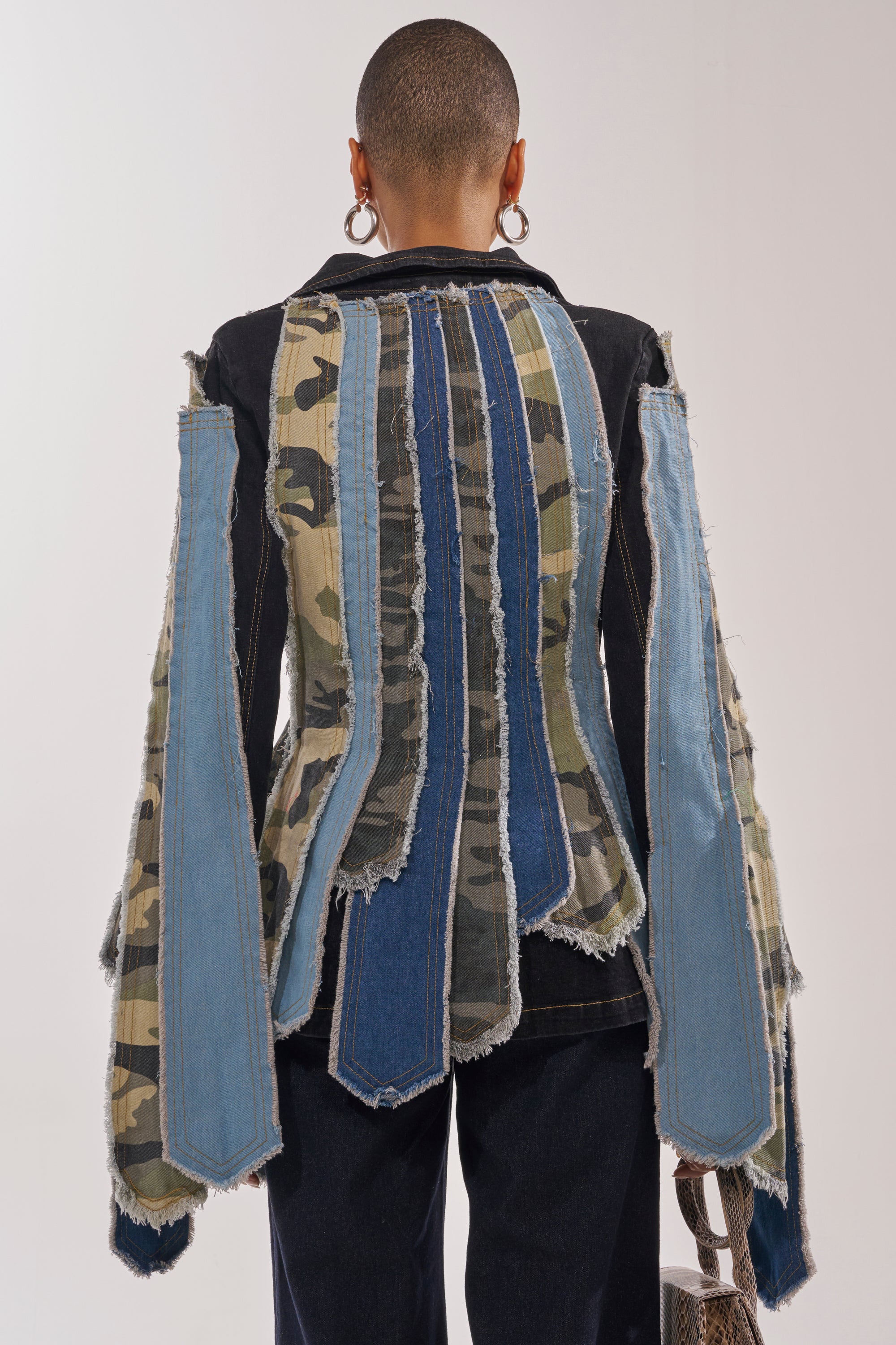 A person with short hair and large hoop earrings stands with their back to the camera, showcasing the HERE FOR IT PATCHWORK BLAZER, which features bold denim and camouflage strips in a striking patchwork design on the back and sleeves. 2
