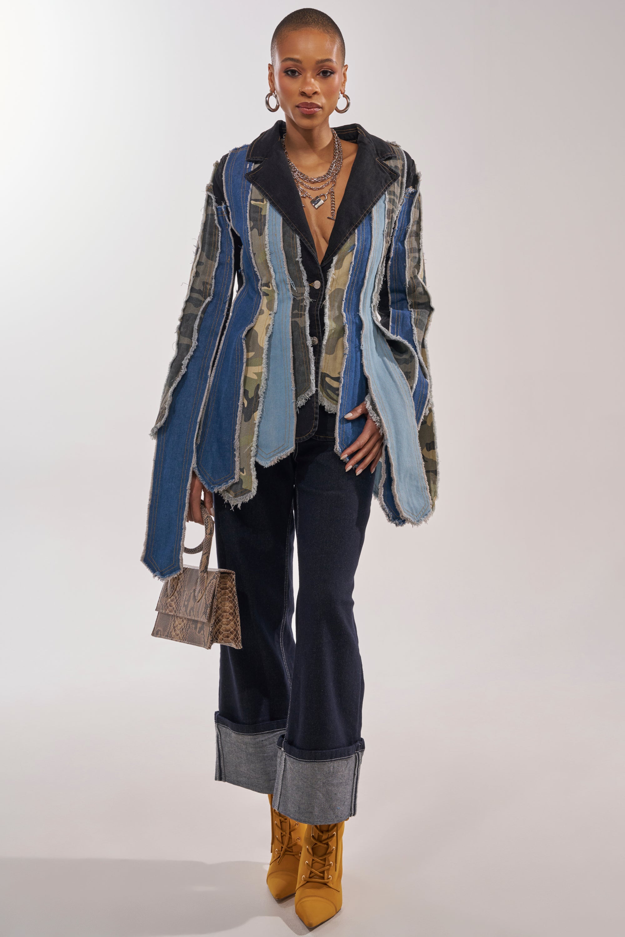 A person stands wearing the HERE FOR IT PATCHWORK BLAZER with frayed edges, wide cuffed dark jeans, mustard yellow boots, layered necklaces, large hoop earrings, and holding a small brown handbag. 3