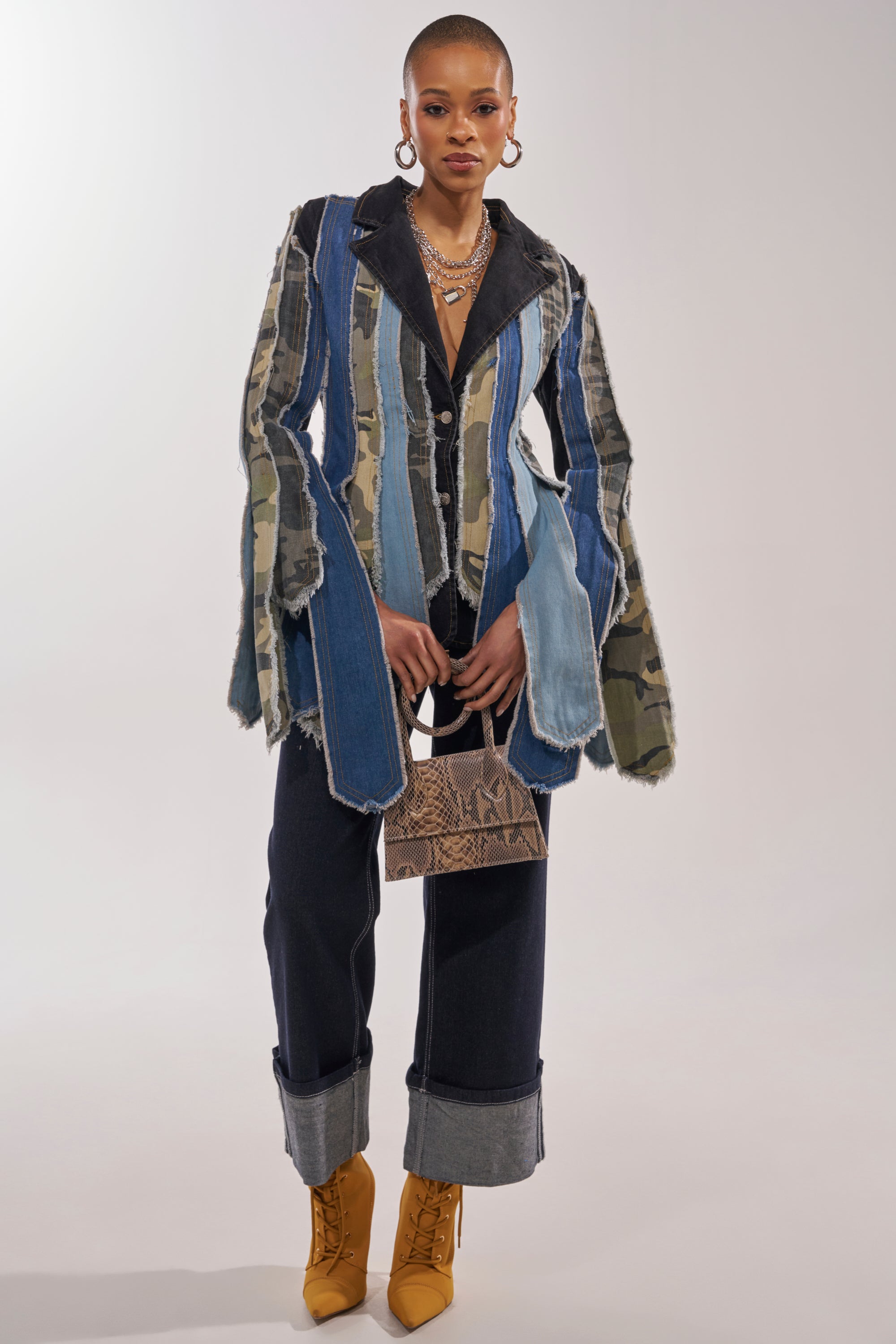 A person stands confidently in the HERE FOR IT PATCHWORK BLAZER with flared sleeves, wide cuffed dark jeans, mustard yellow boots, and a small brown textured handbag. 4