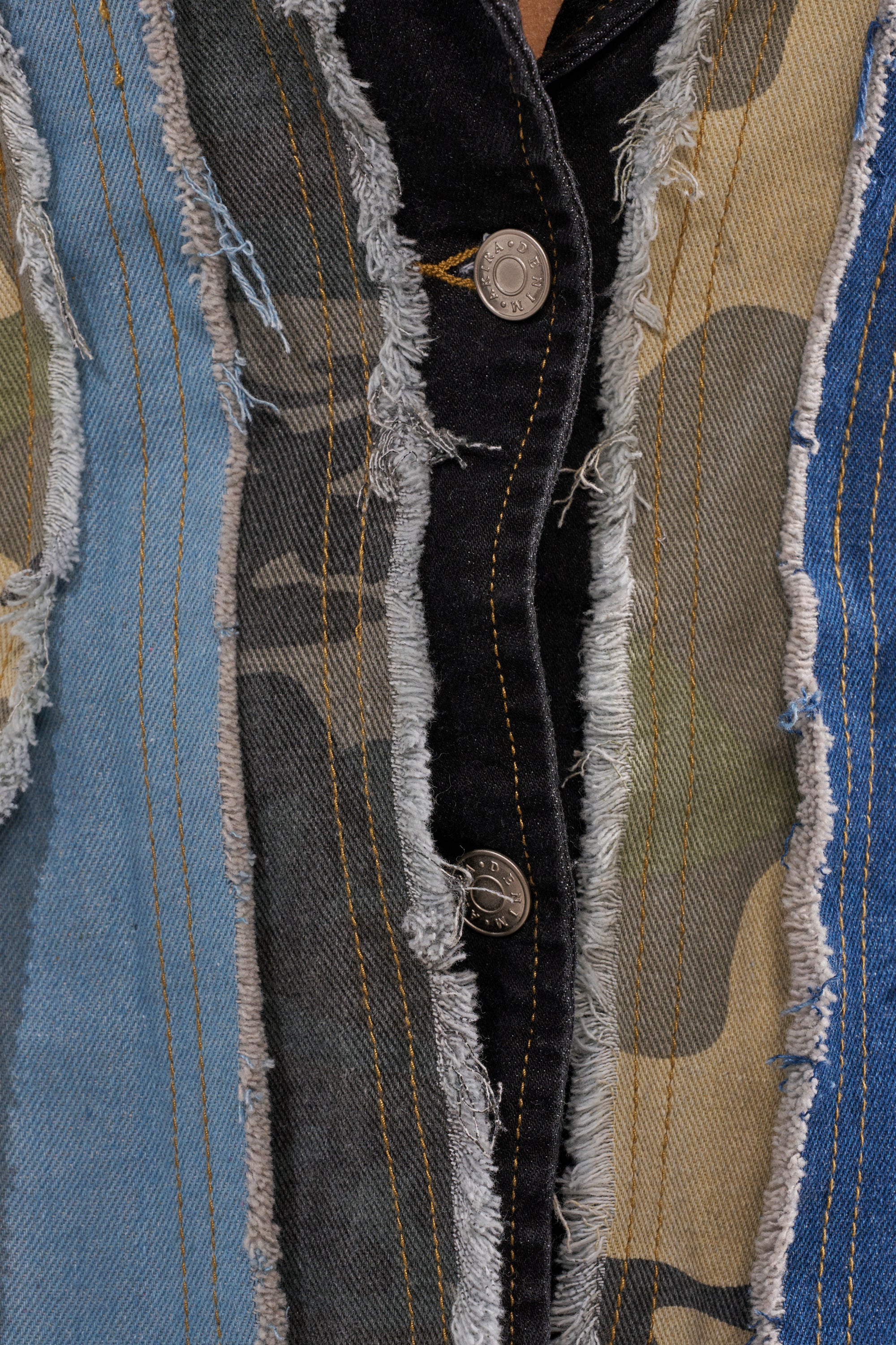 Close-up of frayed denim and camouflage patches with visible seams, stitching, and two metal buttons on dark denim—showcasing the textured style of the HERE FOR IT PATCHWORK BLAZER. 5
