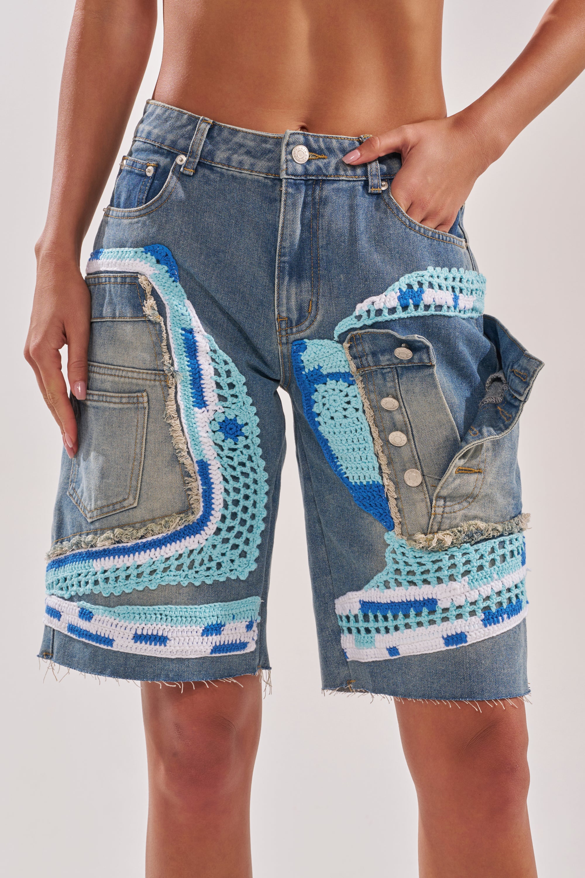 A person wears the GET WITH IT BERMUDA SHORT: high-rise denim with frayed hems, asymmetrical blue, teal, and white crochet knit detailing, distressed edges, and one hand in a pocket. 0