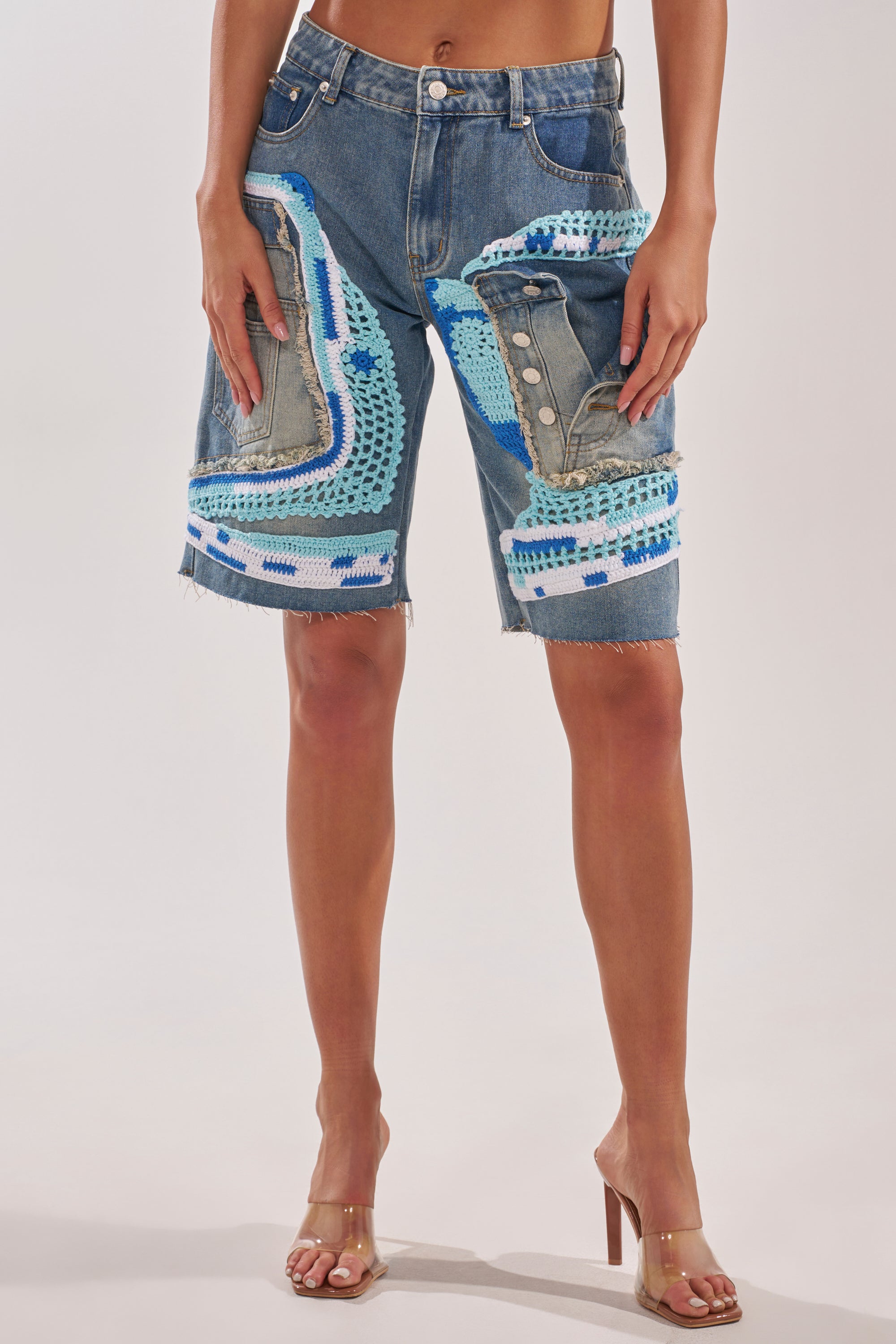 A person wearing the GET WITH IT BERMUDA SHORT, featuring patchwork denim and blue, white, and turquoise crochet knit, paired with clear high-heeled sandals. 1