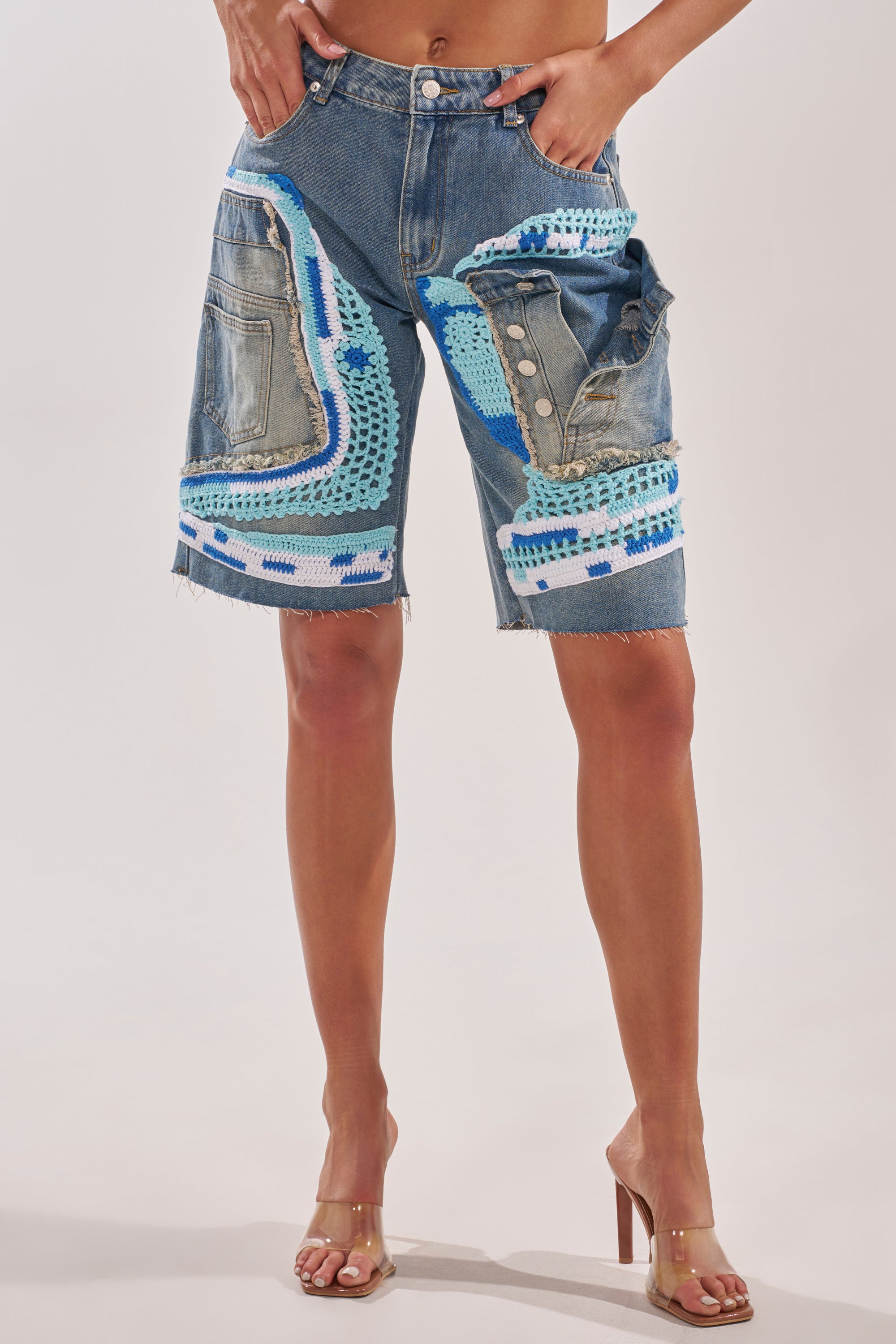 Wearing the GET WITH IT BERMUDA SHORT, featuring high-rise patchwork denim with blue, white, and turquoise crochet patches and frayed hems, paired with clear high-heeled sandals for standout street style. 2