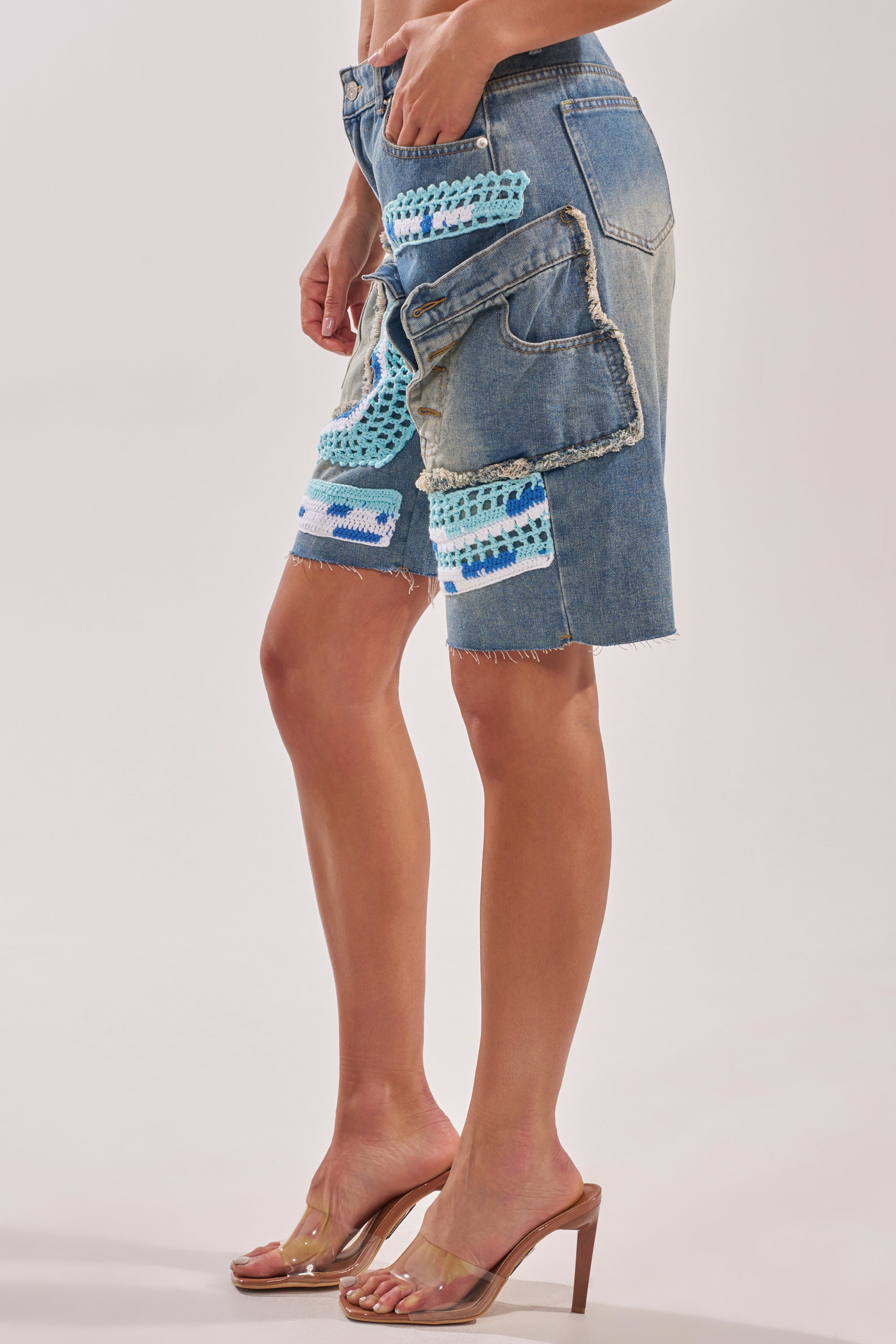 Wearing the GET WITH IT BERMUDA SHORT—high-rise patchwork denim with colorful crochet accents and frayed edges—styled with clear high-heeled sandals for a standout street style look against a plain backdrop. 3