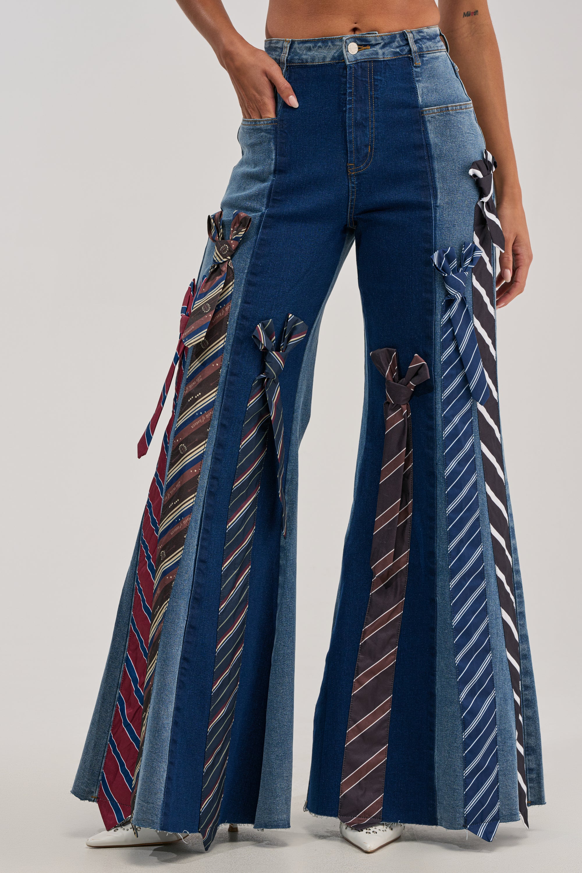A person wearing the TIED TO YOU RELAXED DENIM, a pair of high-waisted, wide-leg jeans crafted from denim in contrasting split wash shades. These jeans are adorned with multiple neckties sewn vertically along the legs, featuring colorful and patterned ties—some with bold plaid details—for a striking and unique look.