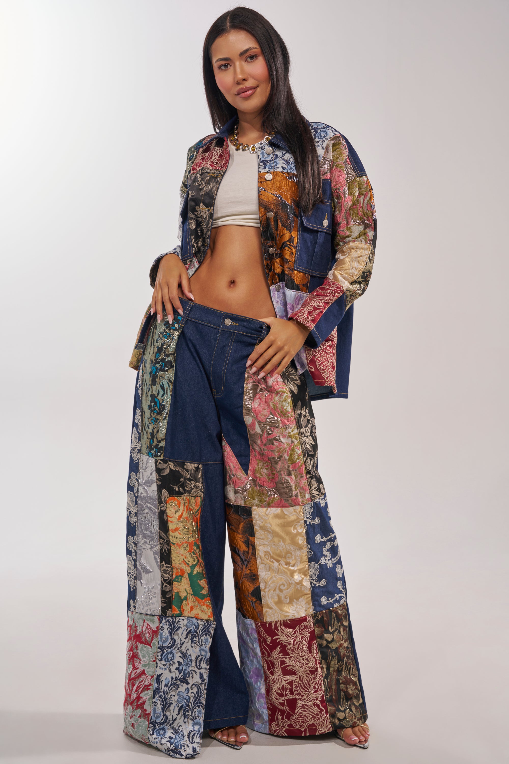 A woman poses confidently in EMOTIONLESS TROUSER wide-leg denim pants with a high-rise waistband, styled with a brocade patchwork denim jacket, white crop top, and necklace against a plain background. 0
