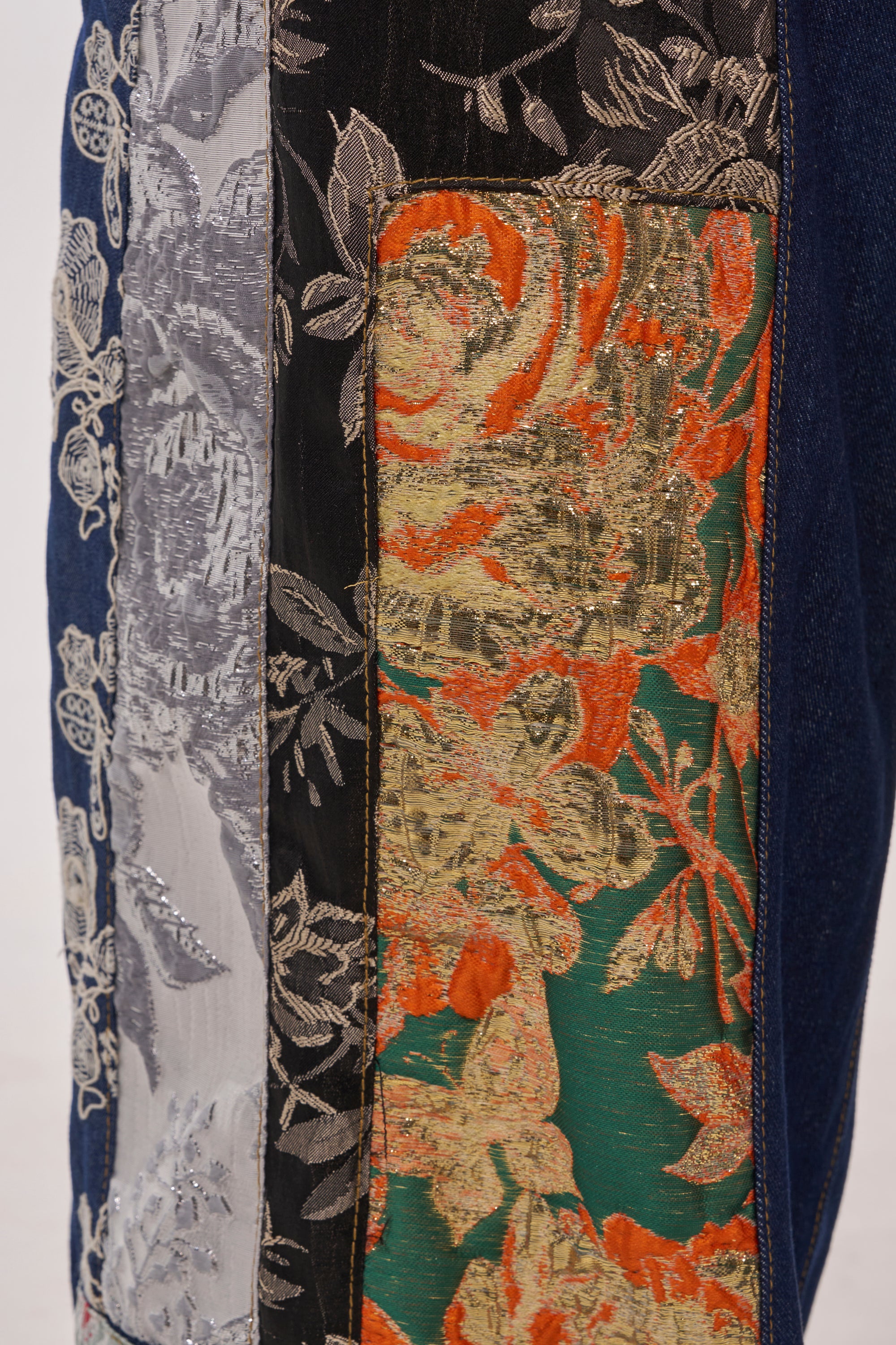 Close-up of brocade patchwork fabric with metallic floral embroidery in silver, gold, orange, and green on black and blue—creating a vibrant texture ideal for the EMOTIONLESS TROUSER or standout accessories. 10