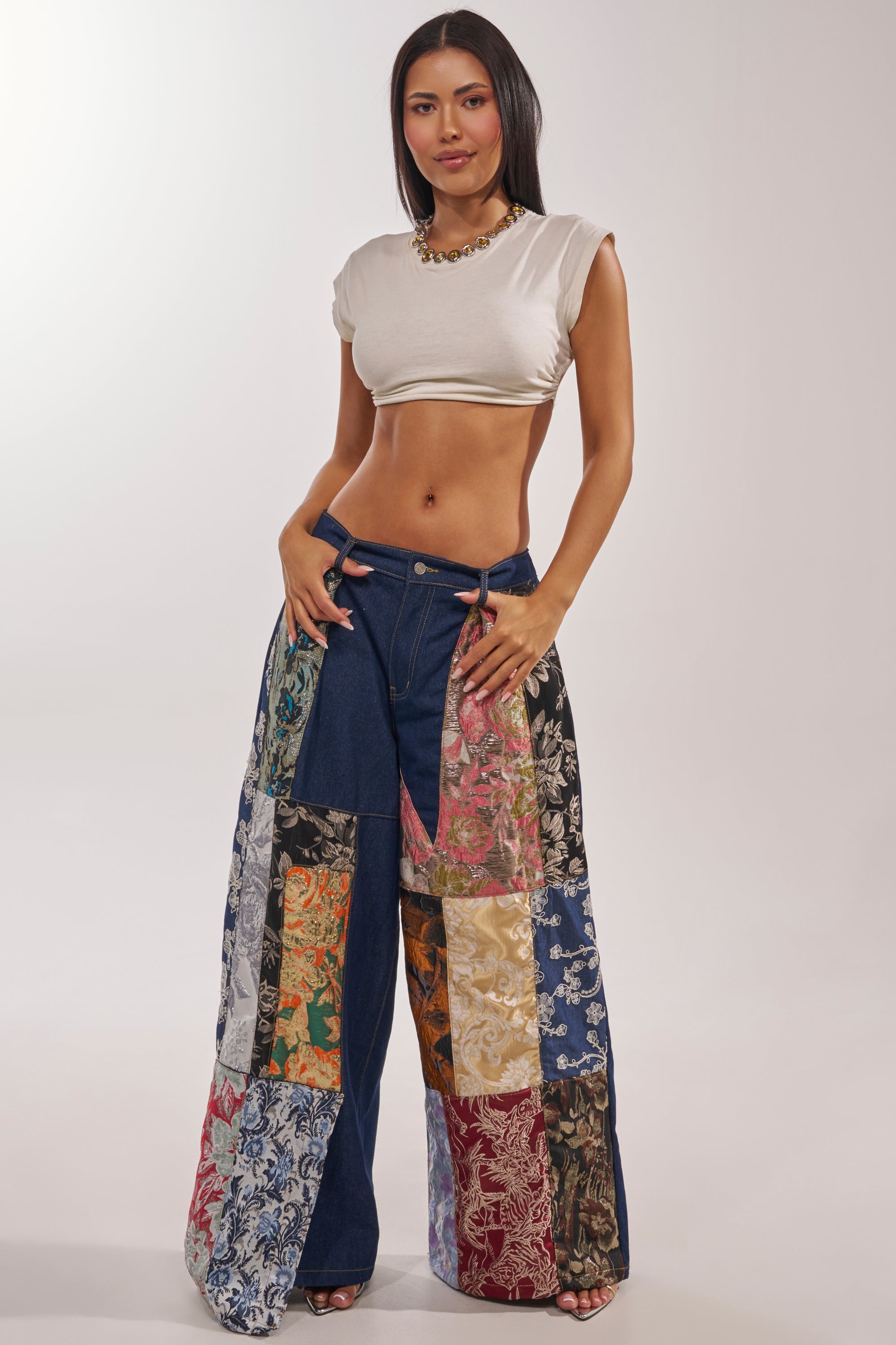 A woman stands confidently in a white cropped top and EMOTIONLESS TROUSER—wide-leg denim with colorful brocade patchwork squares. She has long, straight dark hair against a neutral background. 1