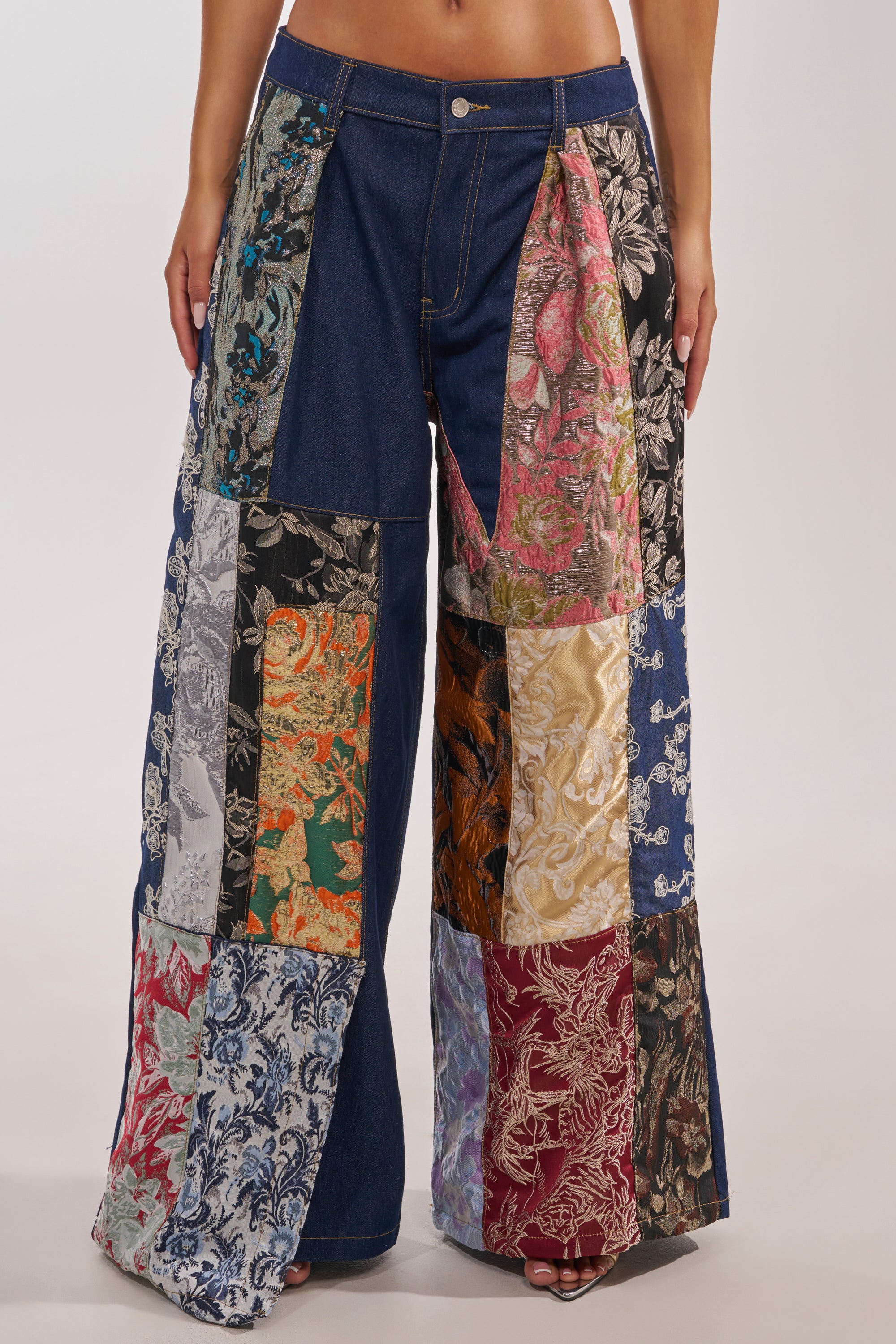 A person wears the EMOTIONLESS TROUSER, wide-leg denim pants with a high-rise waist and vibrant brocade patchwork panels covering both legs from waist to hem. Only the torso to feet are visible. 2