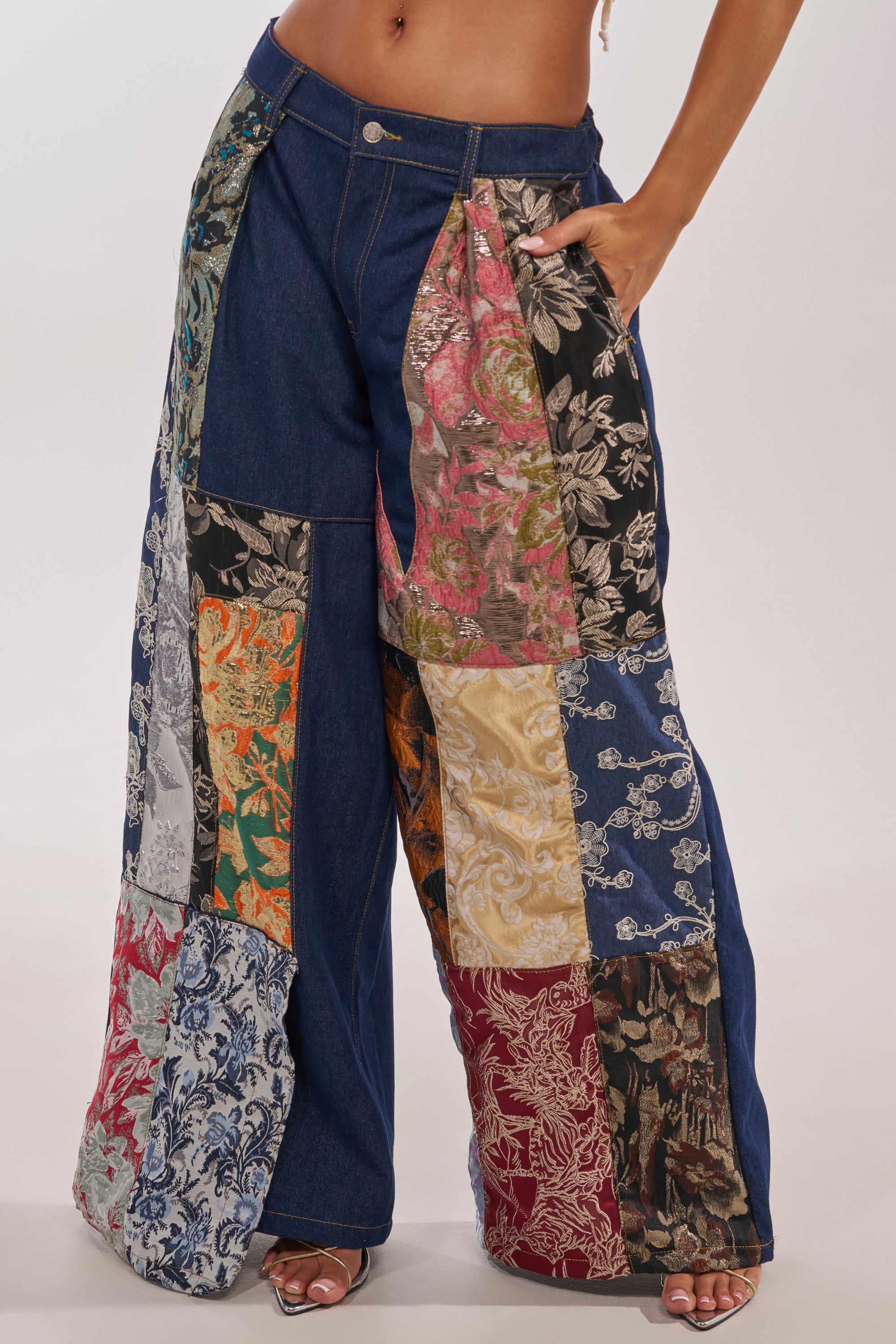 A person wearing the EMOTIONLESS TROUSER, featuring wide-leg denim and large, colorful brocade floral patchwork panels, stands with one hand in their pocket. 3