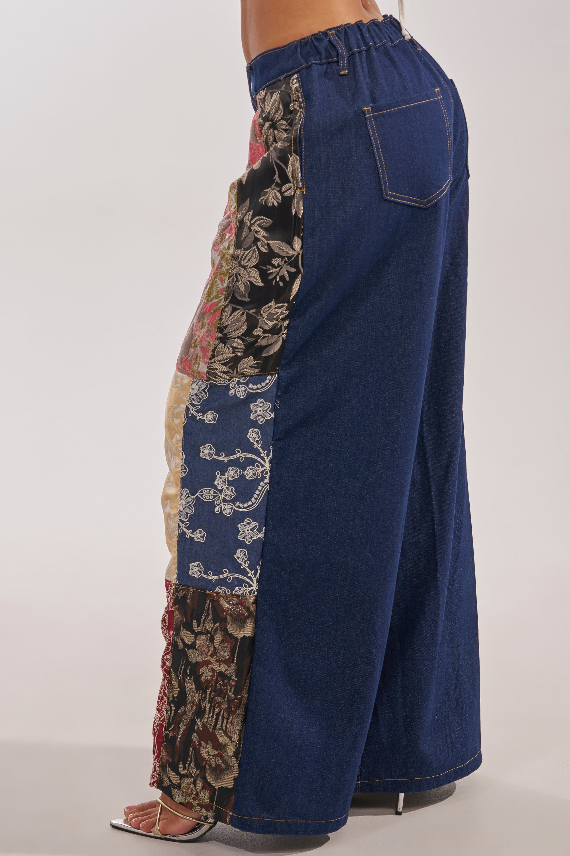 A person wears the EMOTIONLESS TROUSER, featuring wide-leg denim with brocade patchwork side panels of floral and patterned fabrics, a high-rise waistband for comfort, and pairs it with open-toe heeled sandals. 4