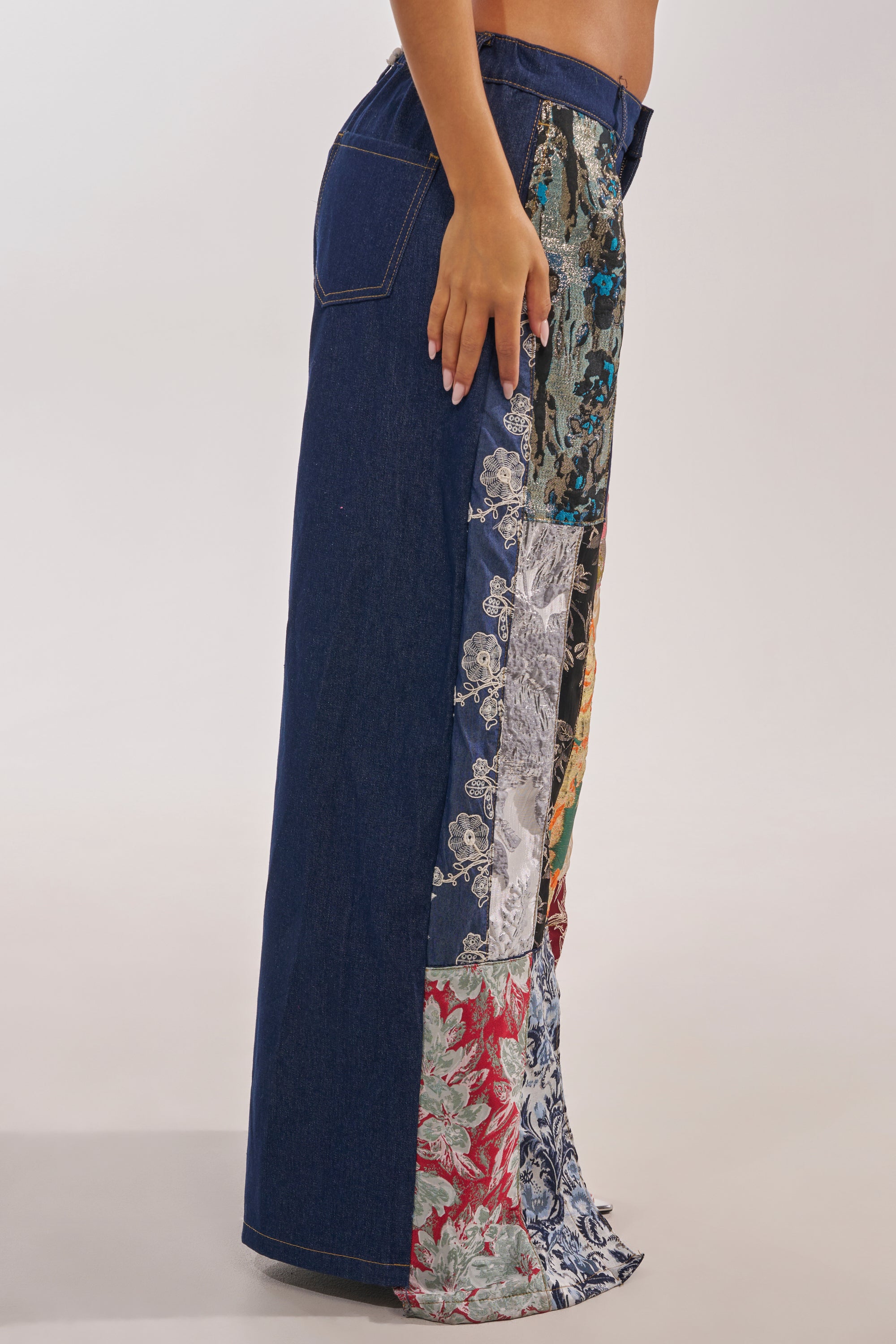 A person stands sideways wearing the EMOTIONLESS TROUSER, a long brocade patchwork skirt crafted from denim and colorful floral fabric panels. 5