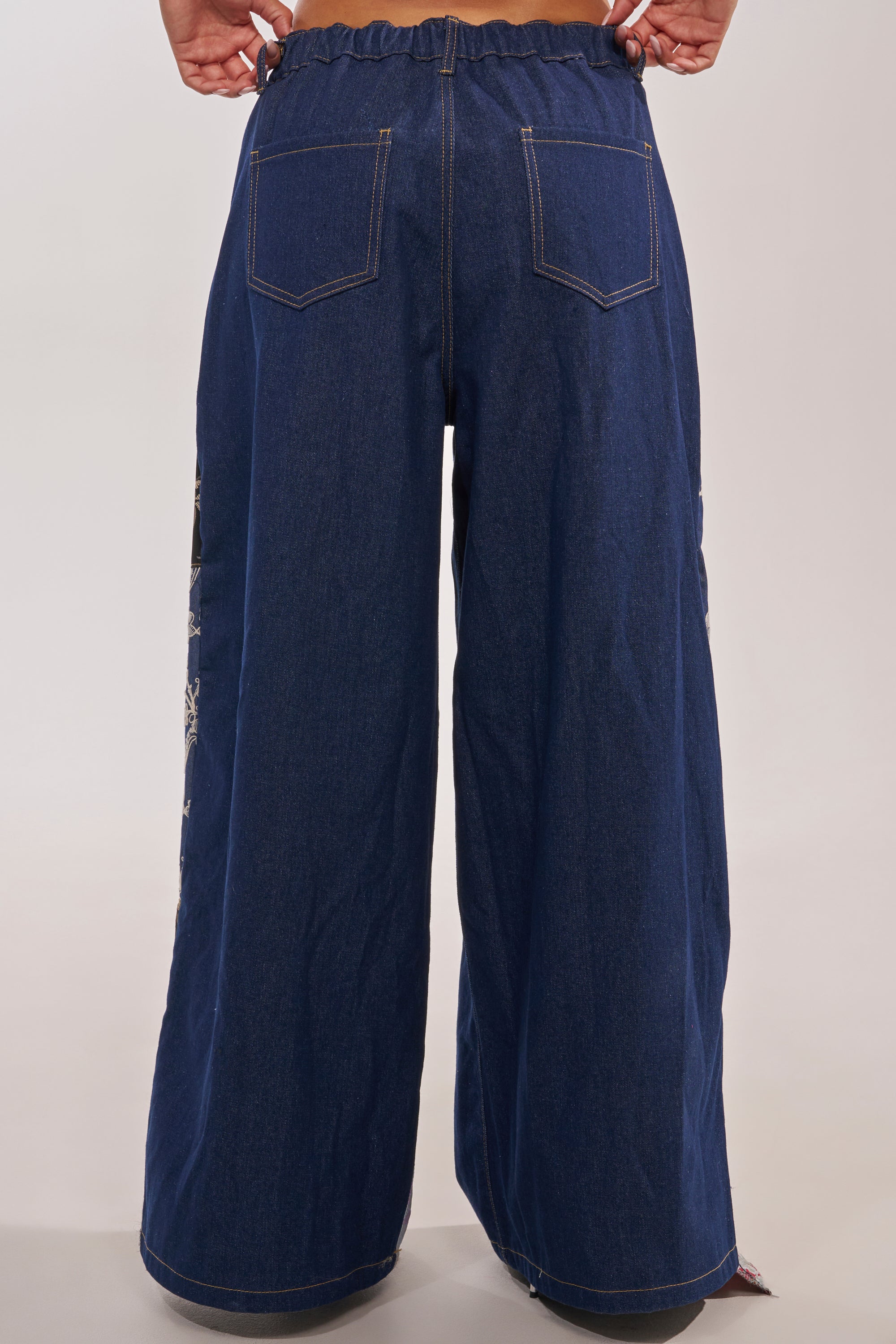 Model showcases the EMOTIONLESS TROUSER—a wide-leg denim with visible back pockets, high-rise waistband, and contrast stitching—standing hands on hips, photographed from behind against a plain light backdrop. 6