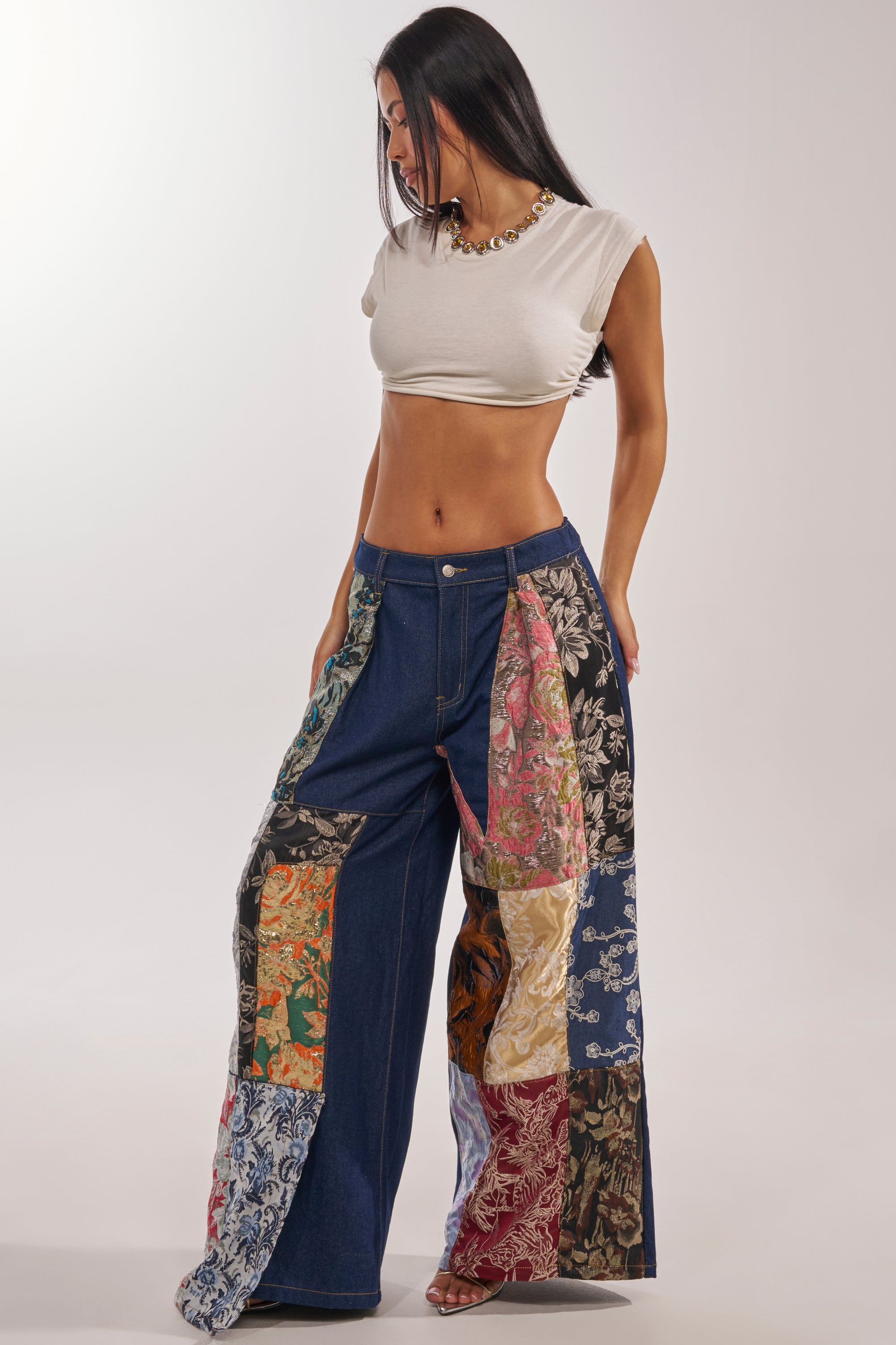 A woman stands against a plain light background wearing a cropped white t-shirt and EMOTIONLESS TROUSER, featuring wide legs, a high-rise waist, and colorful brocade patchwork, looking down to her left. 8