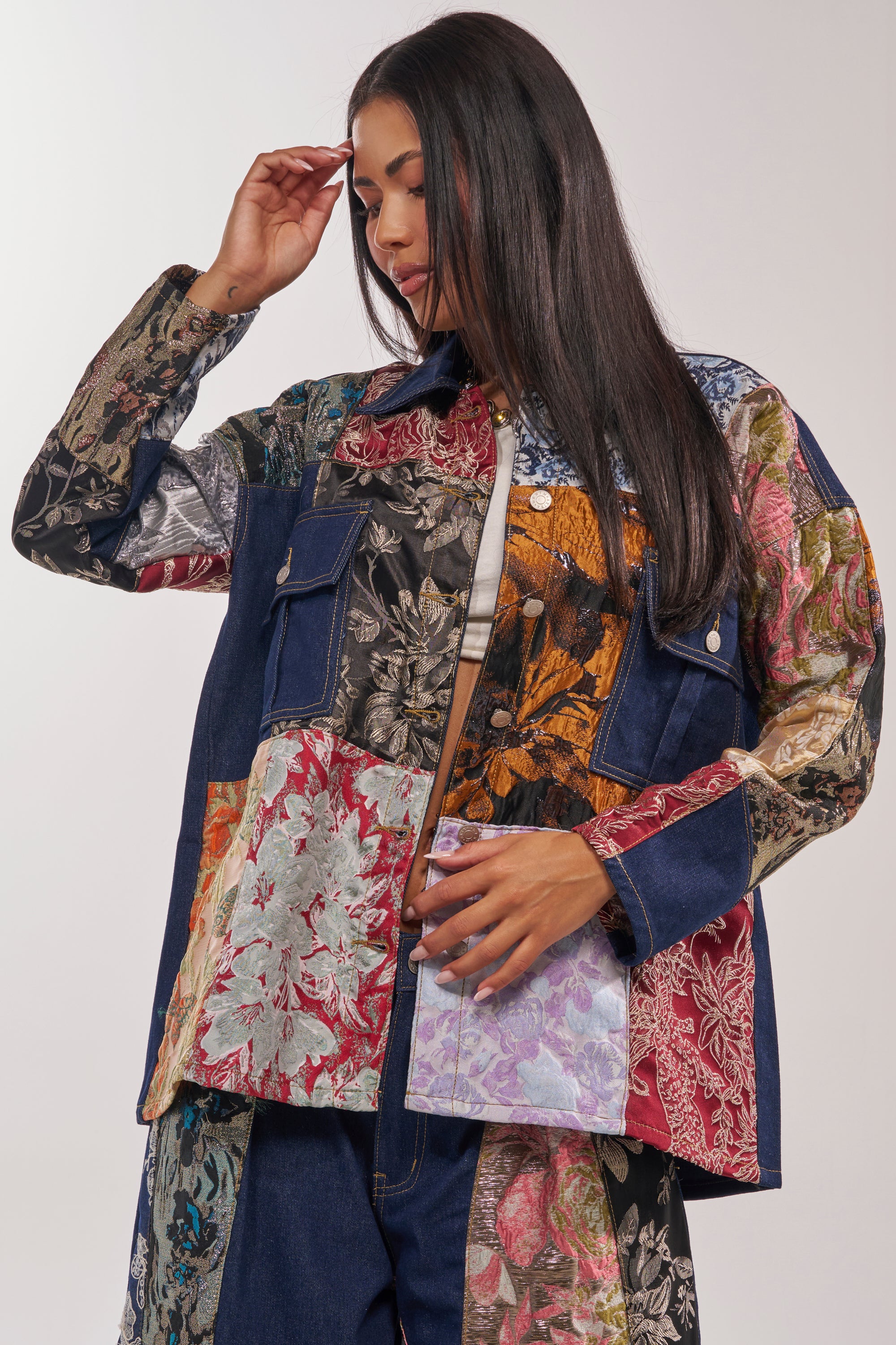 A woman with long dark hair wears the EMOTIONLESS BUTTON DOWN, a loose-fitting denim shirt paired with colorful floral patchwork pants, looking down with one hand near her head and the other resting on her jacket. 0