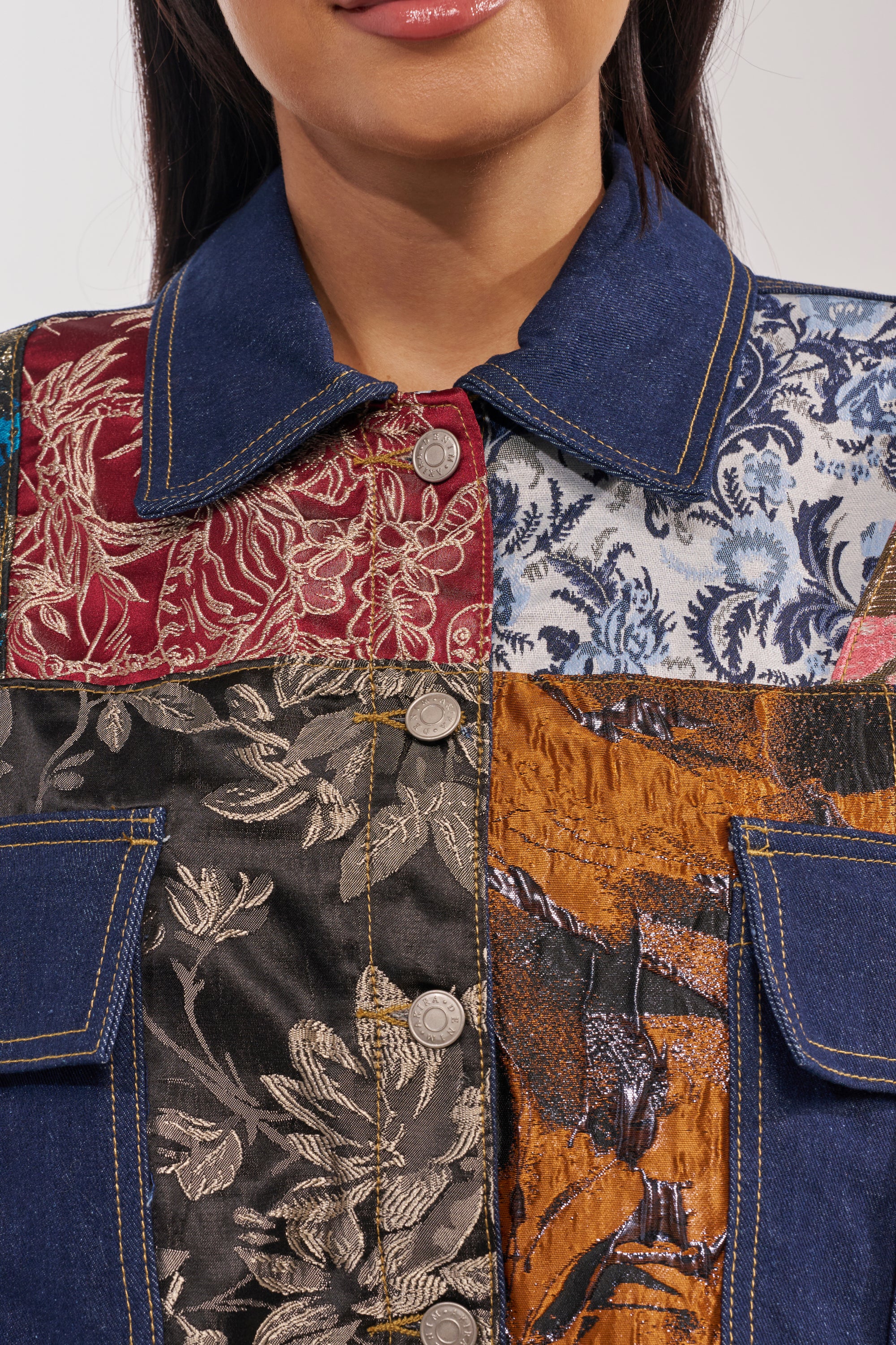 A woman wears the EMOTIONLESS BUTTON DOWN, a patchwork jacket with metallic brocade, denim, floral prints, and utility chest pockets. The photo is cropped to show only her mouth, neck, and upper chest. 9