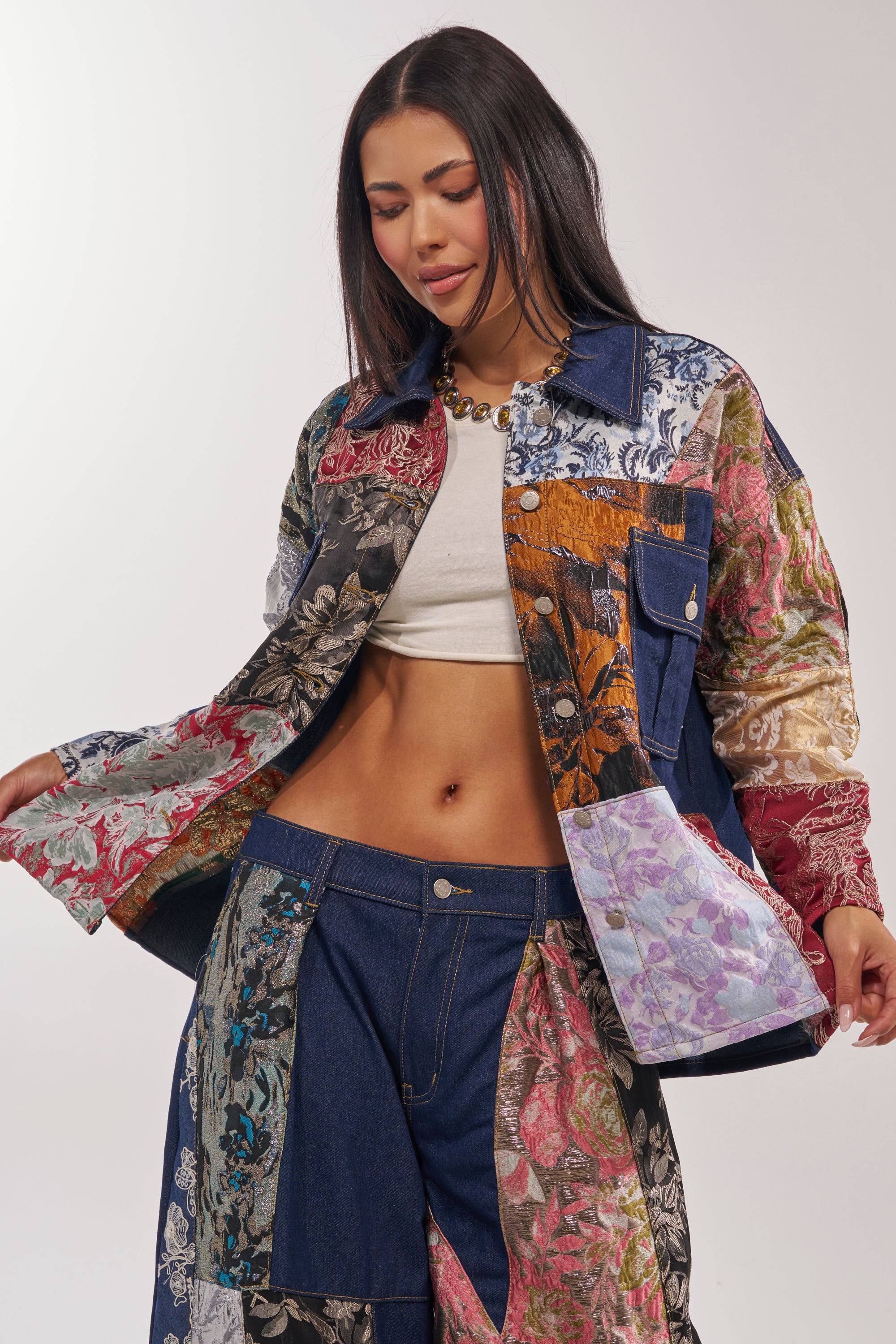 A woman in the EMOTIONLESS BUTTON DOWN—a patchwork denim shirt with floral prints—paired with matching pants, a white crop top, and a chunky necklace, stands against a light background, looking down and smiling slightly. 1