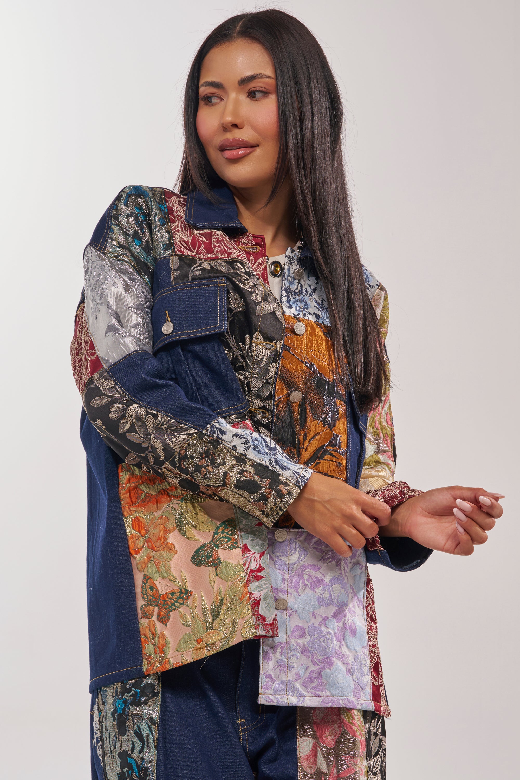 A woman with long dark hair wears the EMOTIONLESS BUTTON DOWN, a colorful patchwork jacket featuring floral designs, denim accents, and metallic brocade. She stands before a plain background, gazing softly to the side with a gentle smile. 2