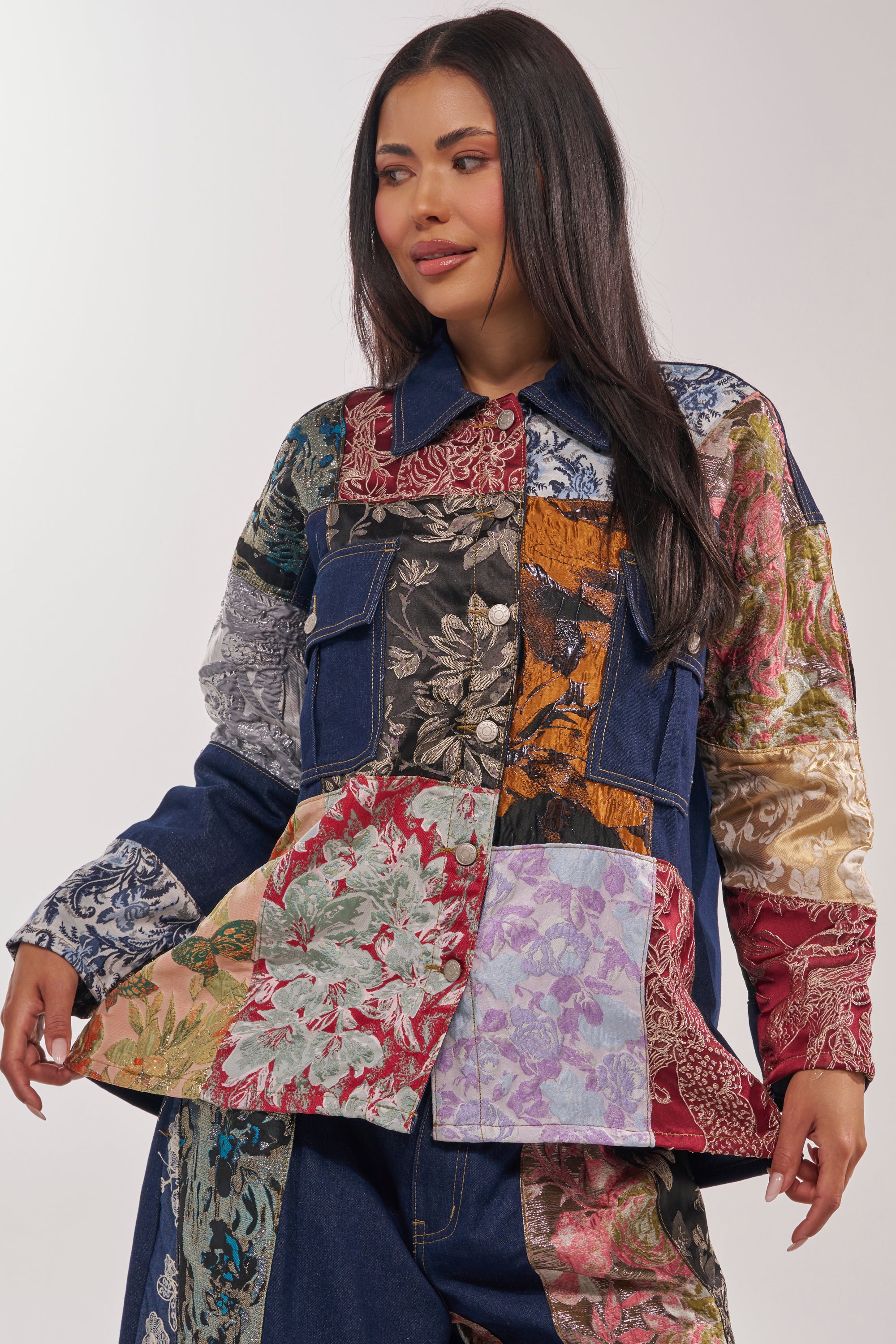 A woman with long dark hair wears the EMOTIONLESS BUTTON DOWN, a patchwork jacket with colorful floral and patterned fabric squares and touches of metallic brocade. She smiles subtly, looking to the side against a plain light background. 3