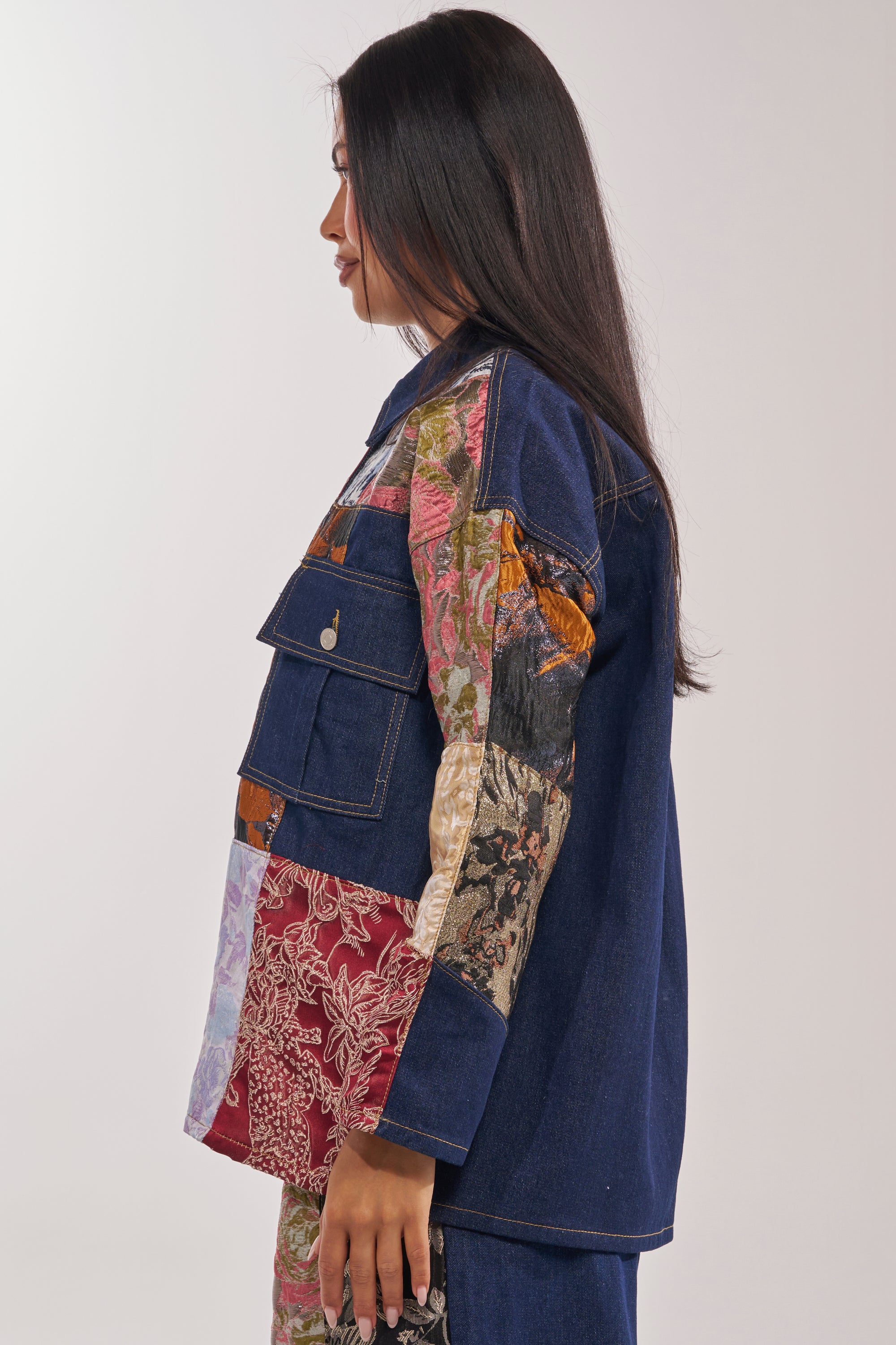 A woman with long dark hair in profile wears the EMOTIONLESS BUTTON DOWN, a patchwork jacket with denim and metallic brocade panels plus utility chest pockets, set against a plain light background. 4