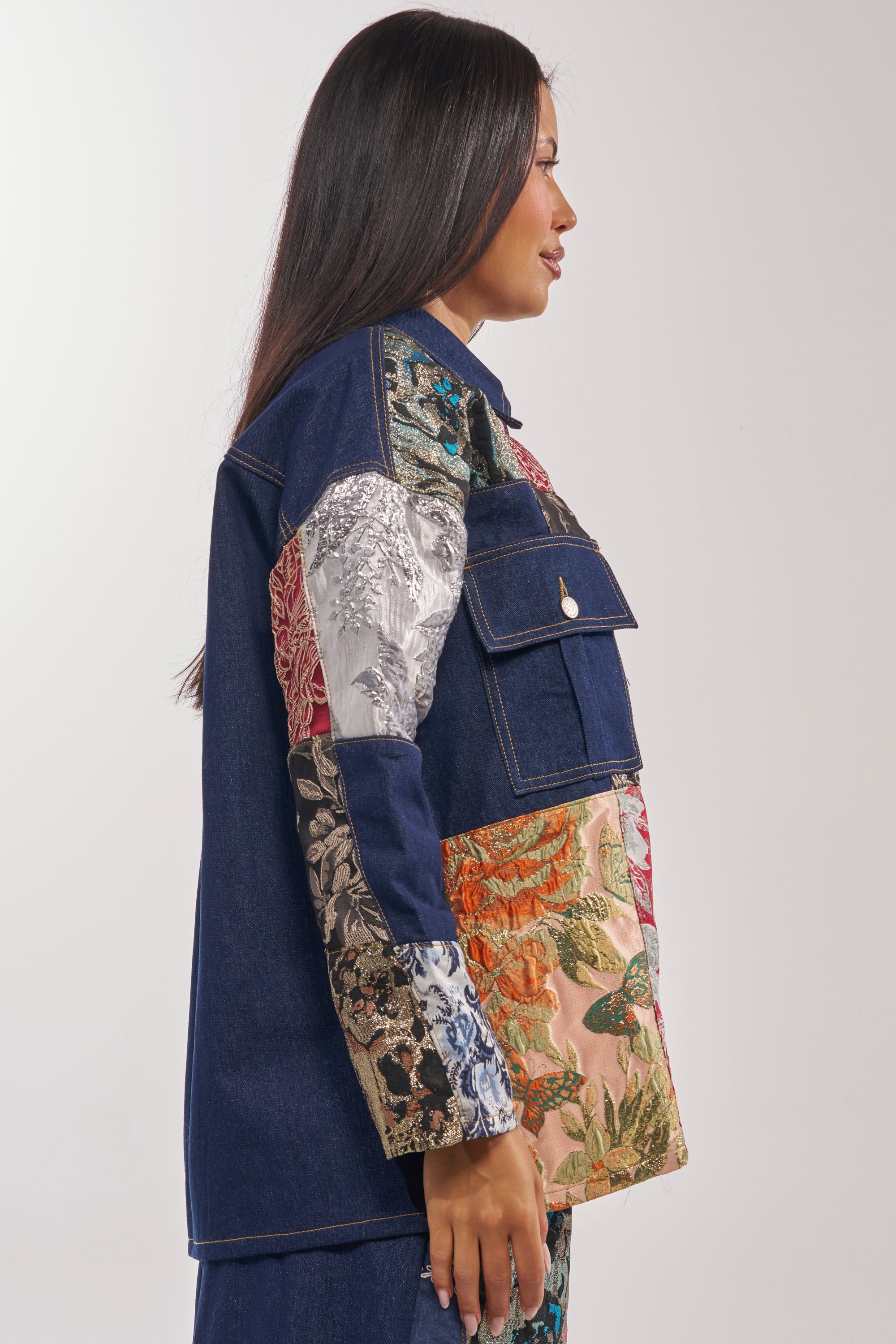 A woman stands in profile wearing the EMOTIONLESS BUTTON DOWN, a denim shirt with utility chest pockets and metallic brocade patchwork featuring colorful floral and metallic designs for a bold, eclectic look. 5