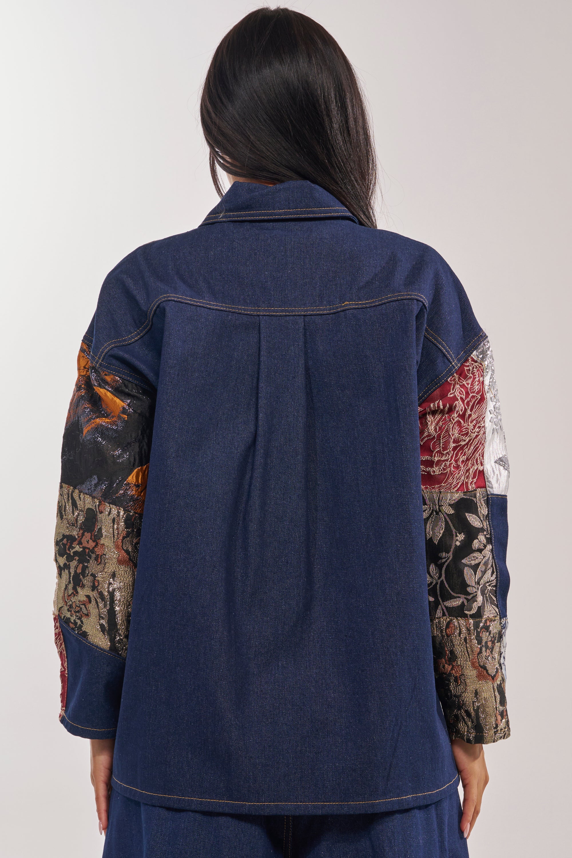 A person with long dark hair is shown from the back wearing the EMOTIONLESS BUTTON DOWN, a blue denim shirt with colorful metallic brocade patchwork on the sleeves, visible stitching, and a relaxed fit. 6
