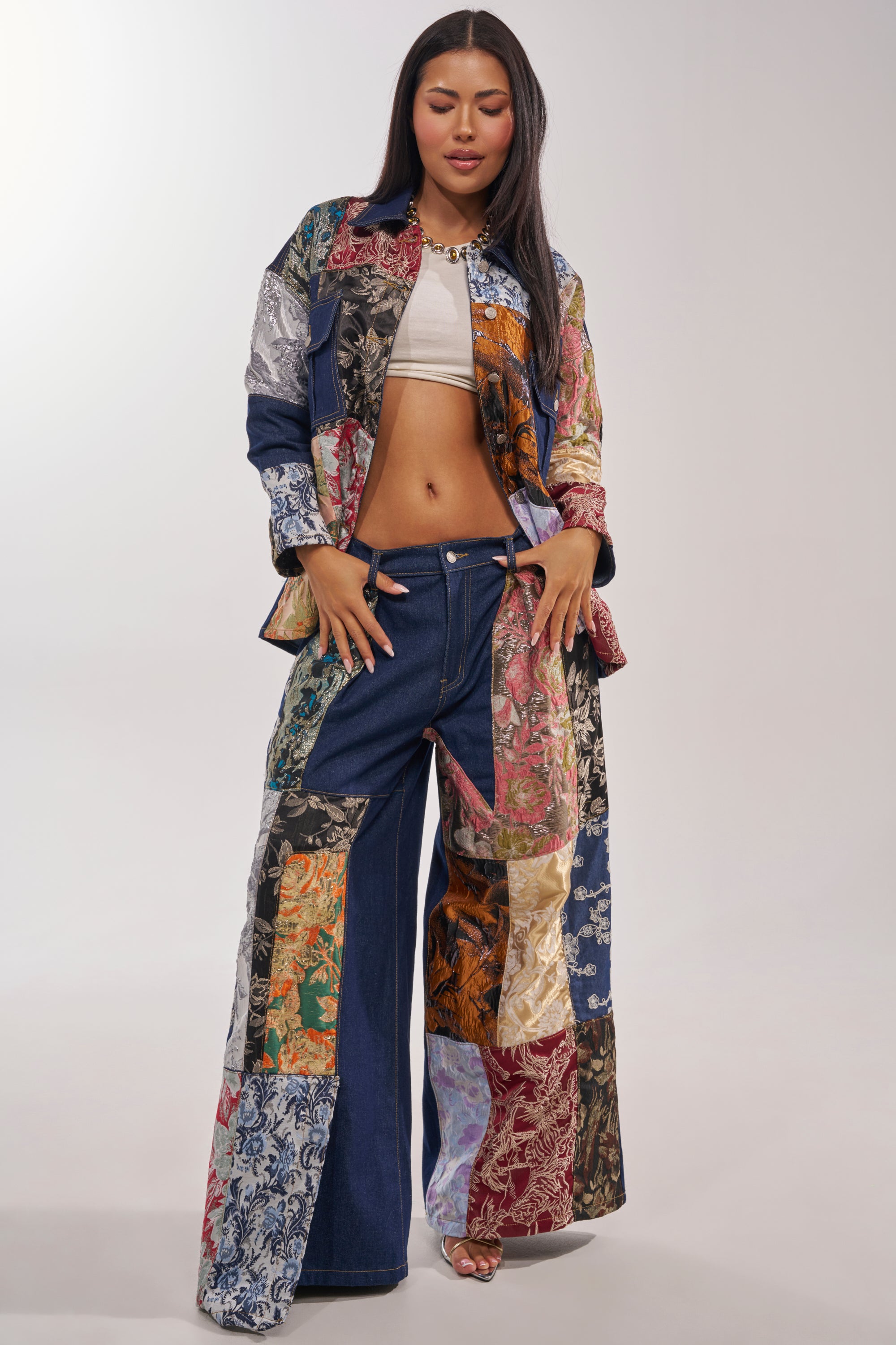 A woman stands confidently in the EMOTIONLESS BUTTON DOWN, a patchwork denim shirt with colorful squares, paired with matching wide-leg pants, a white crop top, and open-toed heels against a plain light background. 8