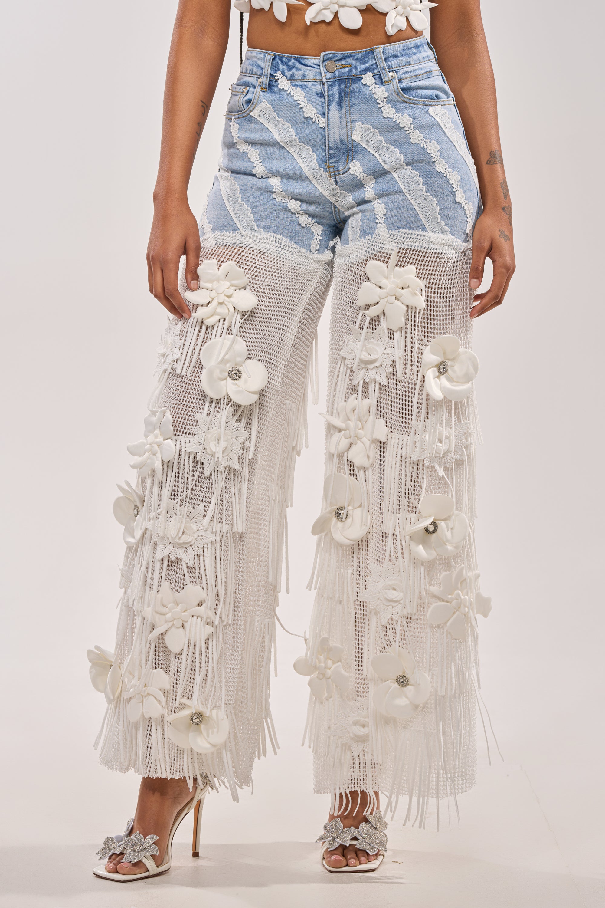 A person wears FESTIVAL GIRL RELAXED DENIM with sheer white mesh overlays featuring large 3D white flowers, paired with floral high-heeled sandals. Only the lower body is shown against a neutral background. 0