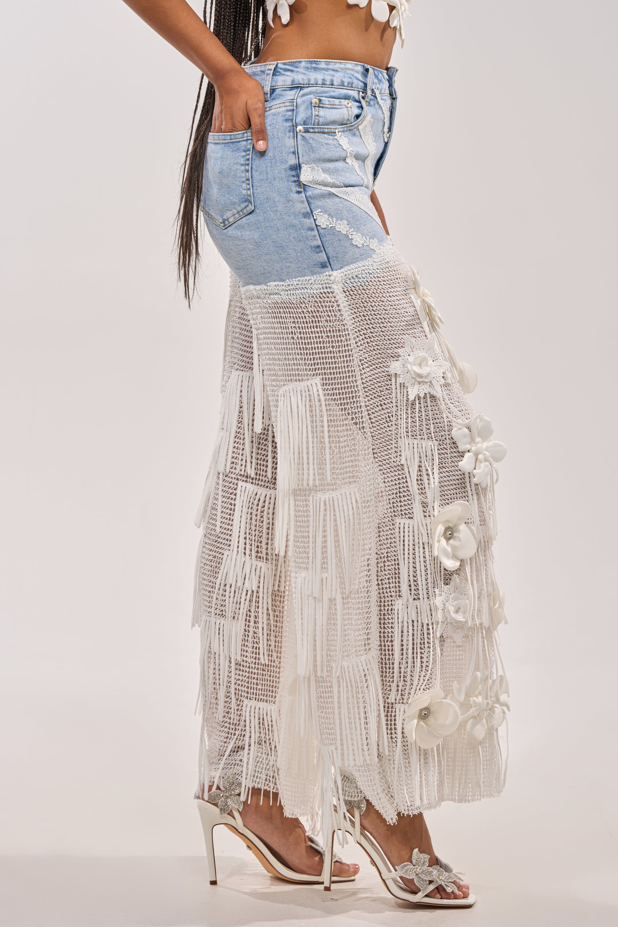 A person wears the FESTIVAL GIRL RELAXED DENIM: unique boho festival pants with denim shorts over a sheer white crochet skirt, detailed with 3D florals and fringe, styled with white heeled sandals and a beaded crop top. 1