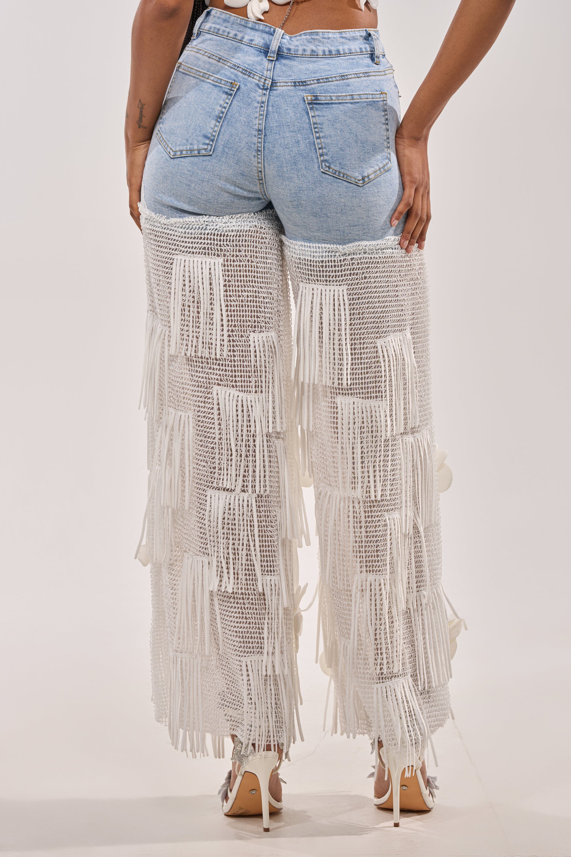 A person is shown from the back wearing FESTIVAL GIRL RELAXED DENIM in light blue crochet with white fringe overlays, paired with high-heeled shoes, against a plain background. 2