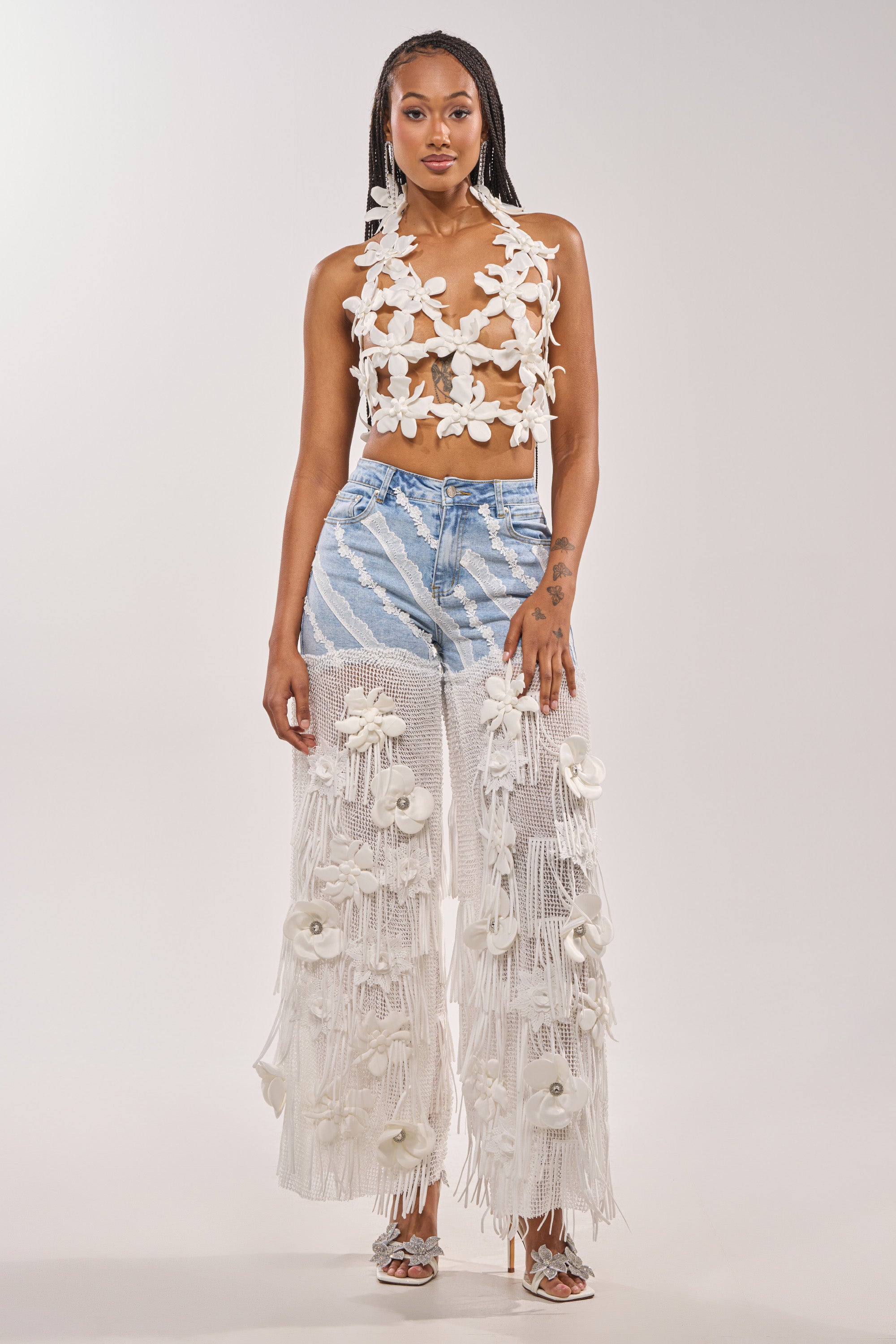 A woman with long braids and strappy heels poses confidently in the FESTIVAL GIRL RELAXED DENIM—crochet fringe pants with sheer, fringed white overlays and 3D flowers—paired with a floral crop top against a plain background. 3