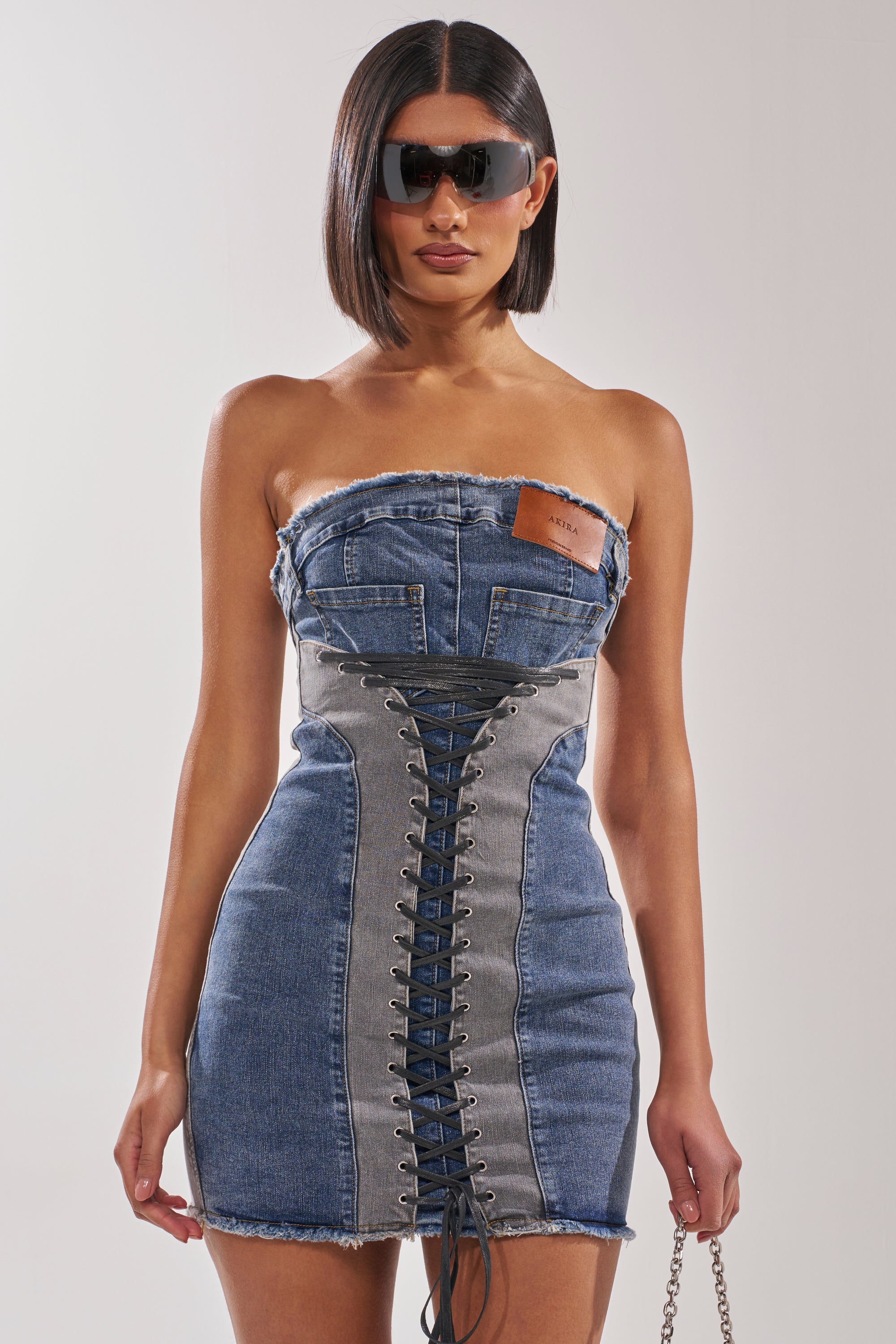 A woman wears the LACED UP MINI DENIM DRESS, a strapless bodycon style with a lace-up front and gray panels. She accessorizes with large black sunglasses, a sleek bob, and a silver chain strap bag. 1