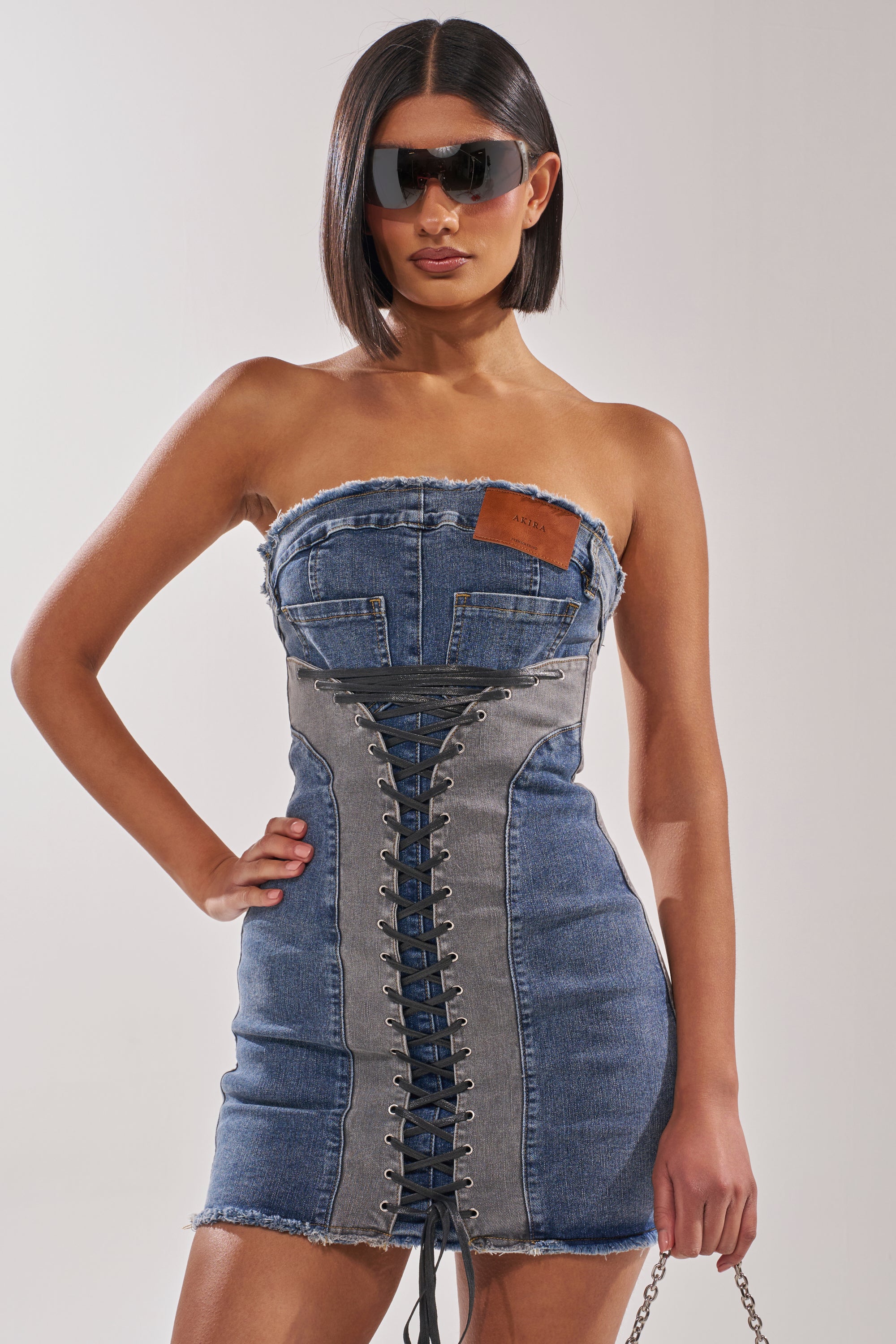 A woman in the LACED UP MINI DENIM DRESS with a gray front panel, sunglasses, and a sleek bob haircut poses with one hand on her hip against a plain background. 2