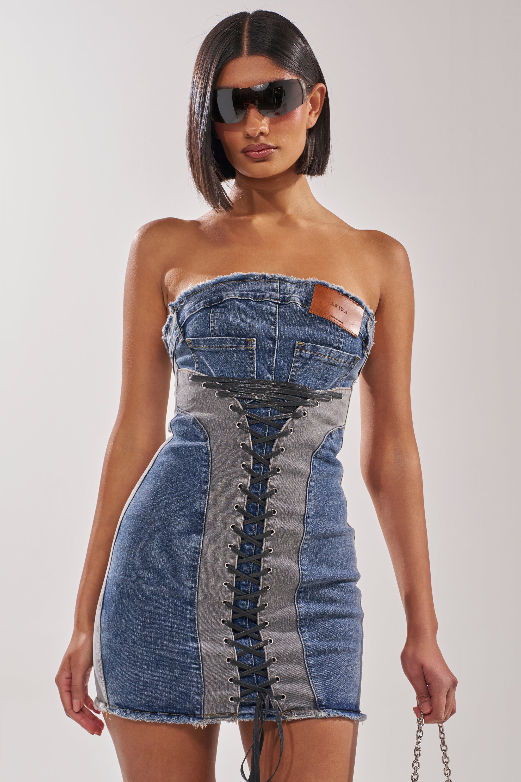 A woman with a sleek bob and dark sunglasses models the LACED UP MINI DENIM DRESS, a strapless, bodycon mini with lace-up front and leather patch detail, standing against a plain background. 3