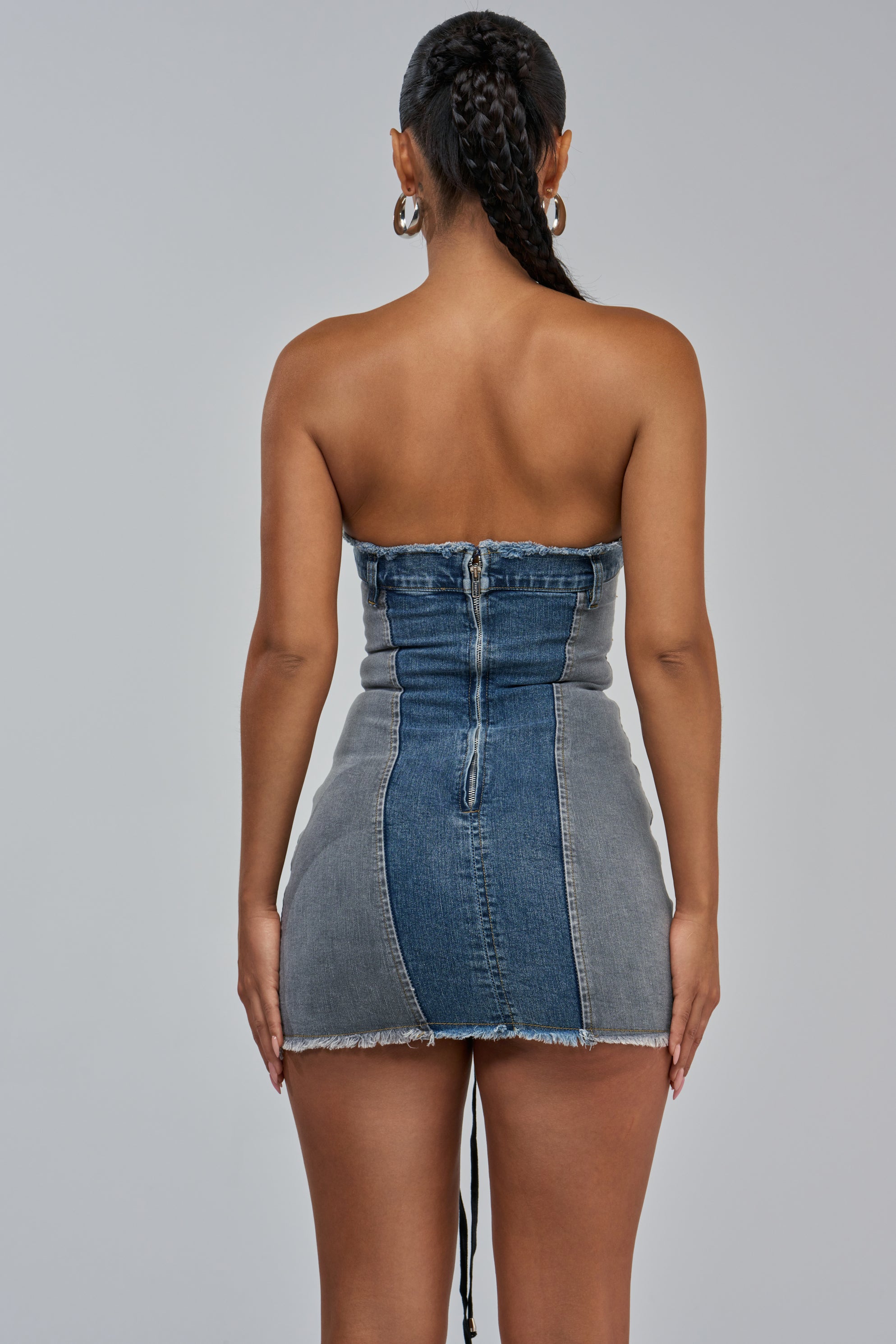 A woman with braided hair is seen from the back against a plain, light gray background, wearing the LACED UP MINI DENIM DRESS—a strapless, form-fitting bodycon mini dress with a frayed hem that makes for an effortlessly cool streetwear concert outfit.