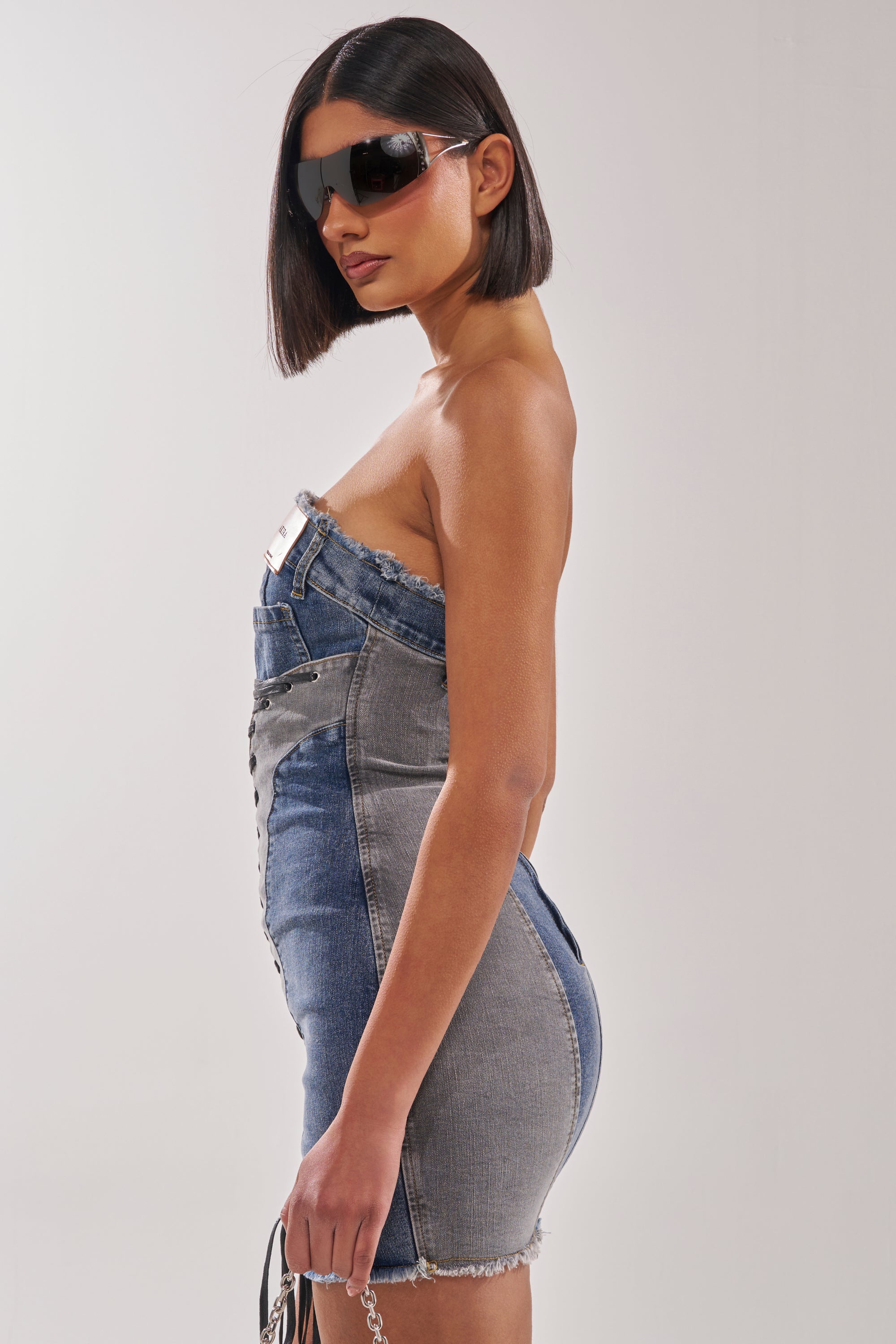 A woman with a sleek bob wears oversized black sunglasses and the LACED UP MINI DENIM DRESS, a strapless, form-fitting mini bodycon style. She carries a small handbag and stands sideways against a plain white background. 4