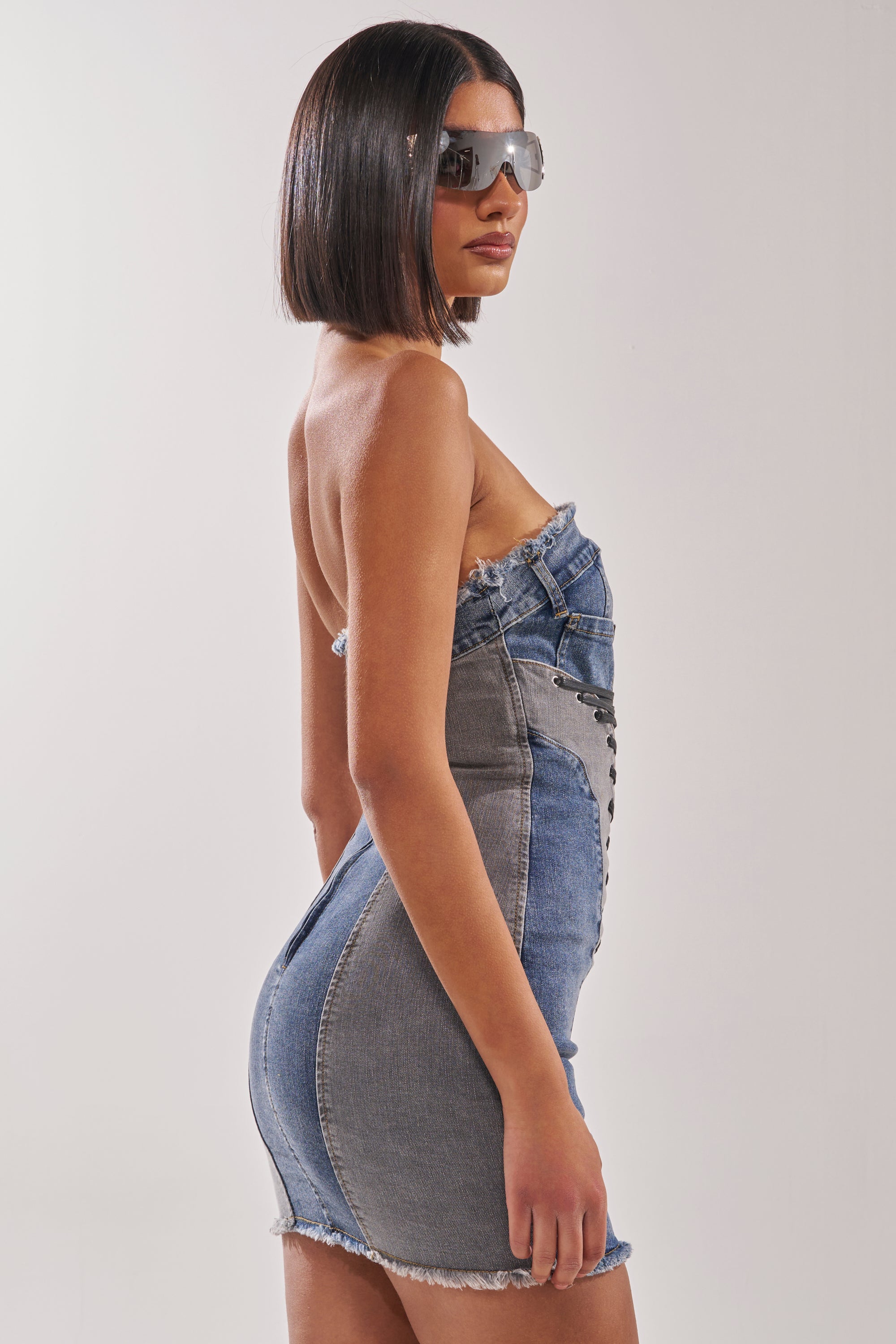A woman with a sleek bob haircut and large sunglasses stands in profile against a white background, modeling the LACED UP MINI DENIM DRESS. 5