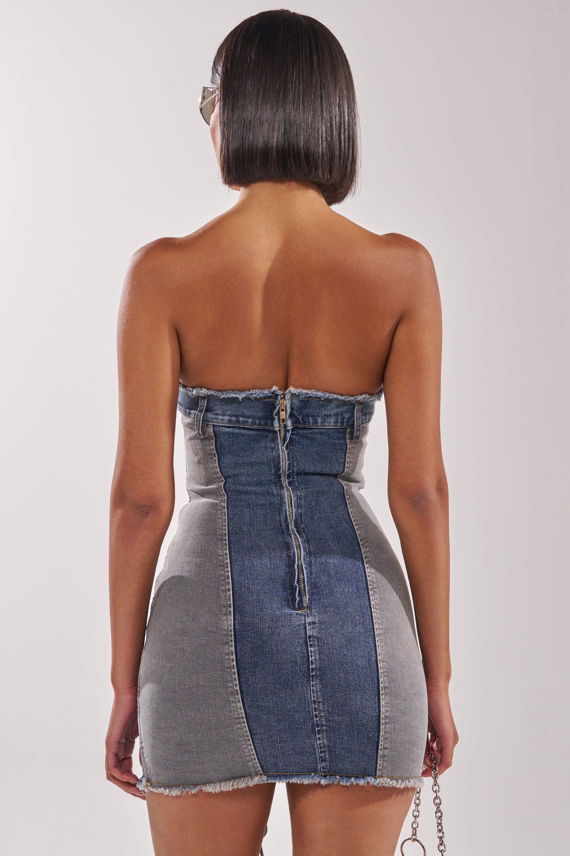 A woman with a short bob is seen from behind wearing the LACED UP MINI DENIM DRESS—a strapless, bodycon mini dress with a visible back zipper and frayed hem—holding a chain-strap purse in her right hand. 6