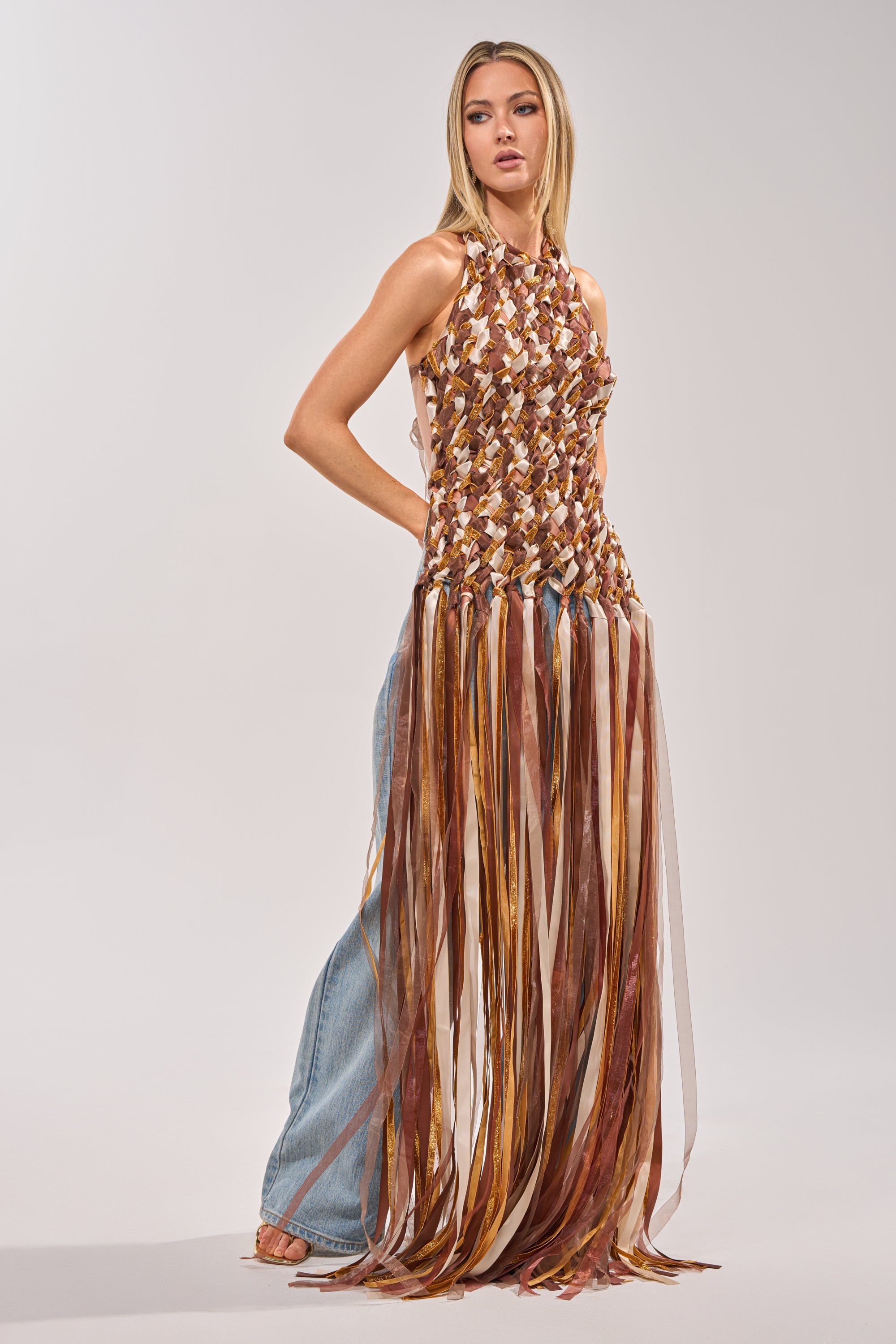 A woman wears the FESTIVAL GIRLY TOP—a fringe statement piece with long, multicolored ribbons—paired with light blue jeans and sandals. She gazes to the side against a simple background. 1