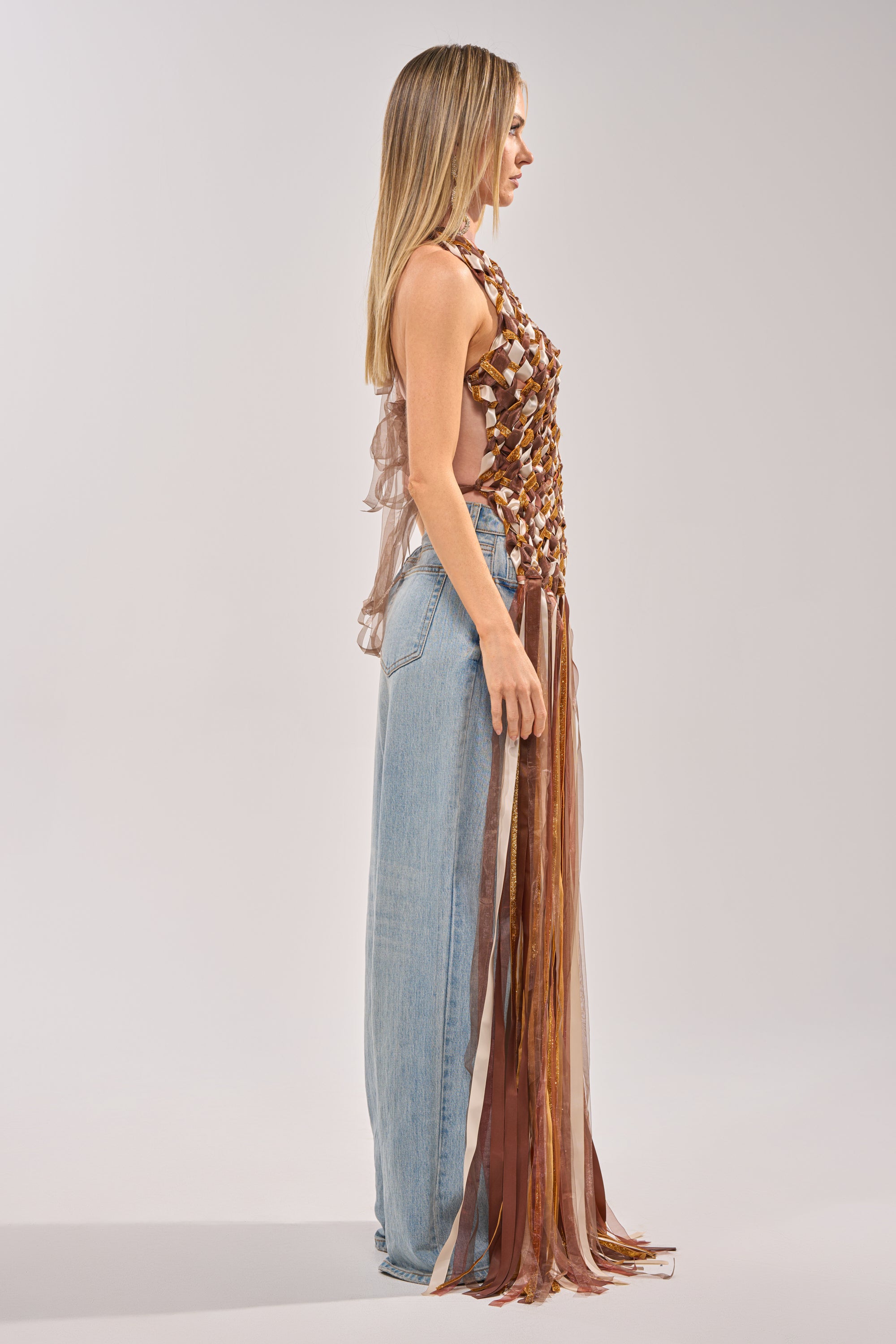 A woman stands in profile against a plain light gray background wearing loose, light blue jeans and the FESTIVAL GIRLY TOP—a sheer, sleeveless halter top featuring intricate design and long, flowing brown and tan woven ribbons. 3