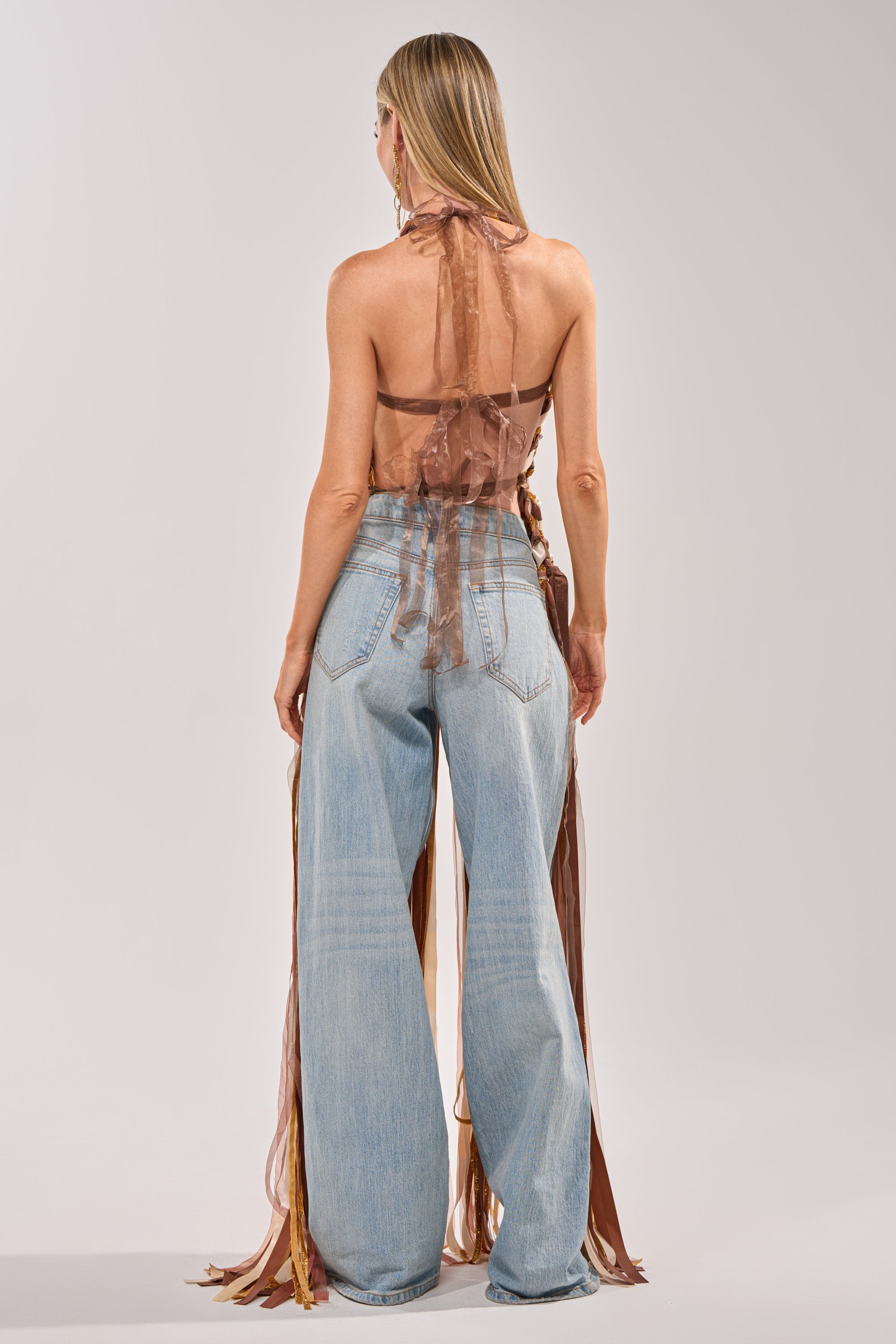 A woman, seen from behind, wears the FESTIVAL GIRLY TOP—a neutral ribbon halter with long ties—paired with loose light blue jeans. Her straight blonde hair drapes down her back against a plain, light backdrop. 4