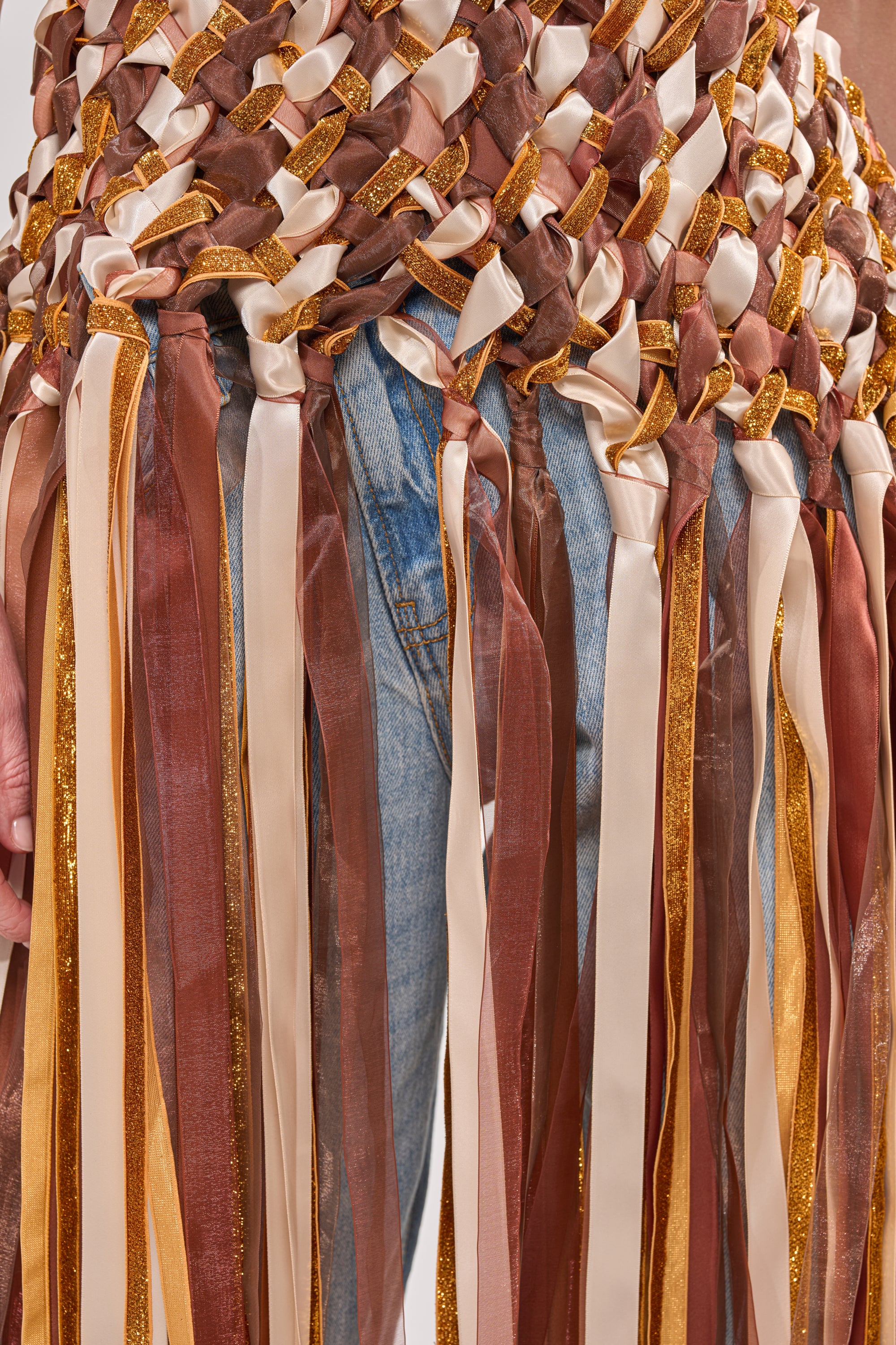 Close-up of a person in blue jeans wearing the FESTIVAL GIRLY TOP—a woven halter featuring interlaced brown, gold, cream, and beige ribbons with long fringe strands—an eye-catching statement piece for festival fashion. 5