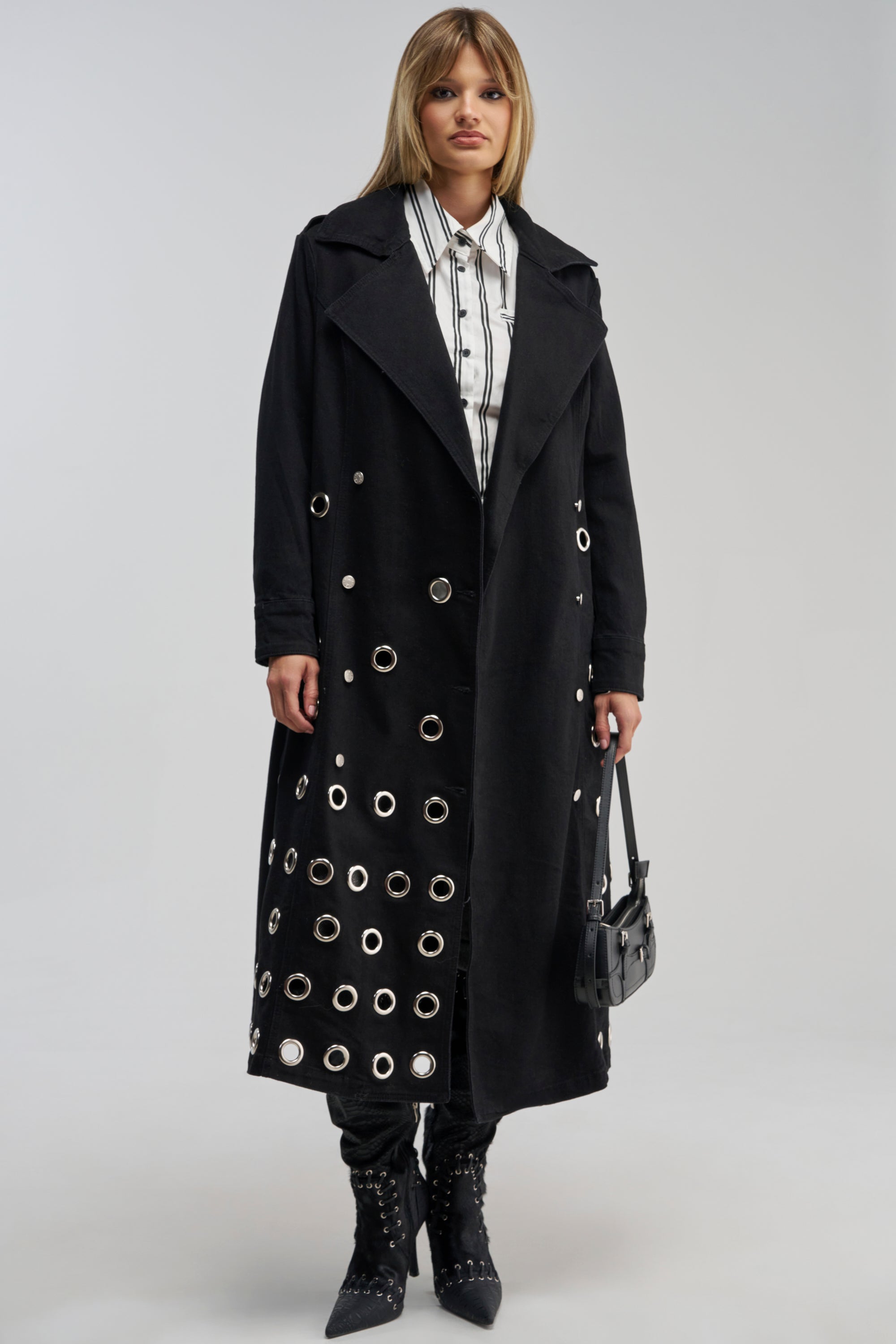 A woman stands wearing the PHOENIX DENIM TRENCH, a striking black denim trench coat adorned with silver grommets, layered over a white button-up shirt with black stripes, paired with black pants, pointed boots, and holding a small black handbag—capturing the essence of modern streetwear.