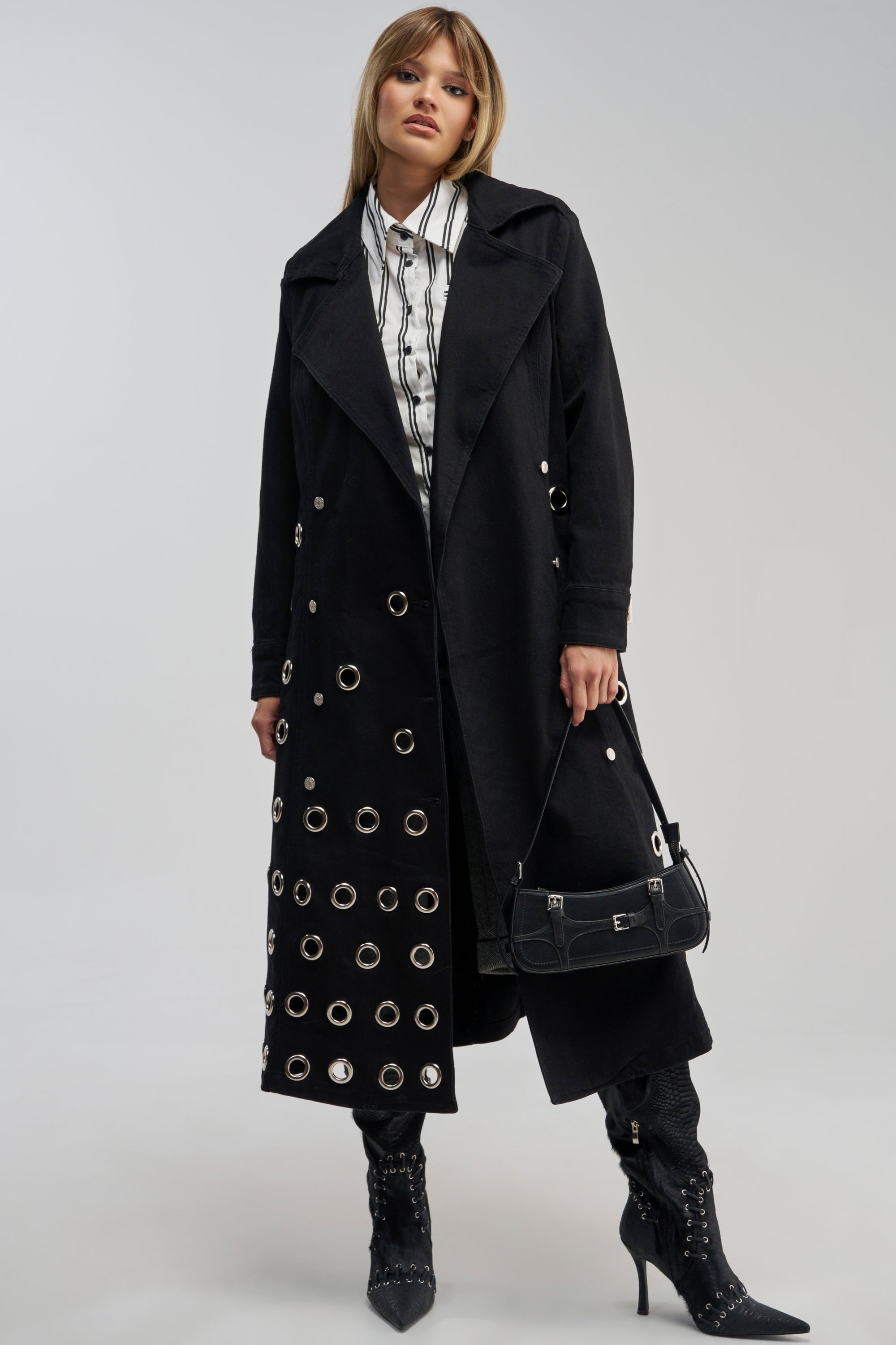 Model wears the PHOENIX DENIM TRENCH, a long black denim coat featuring silver grommet details, paired with black stiletto boots, a crisp striped white shirt, and a small black handbag. She stands confidently against a plain light background, perfectly capturing sleek streetwear style.