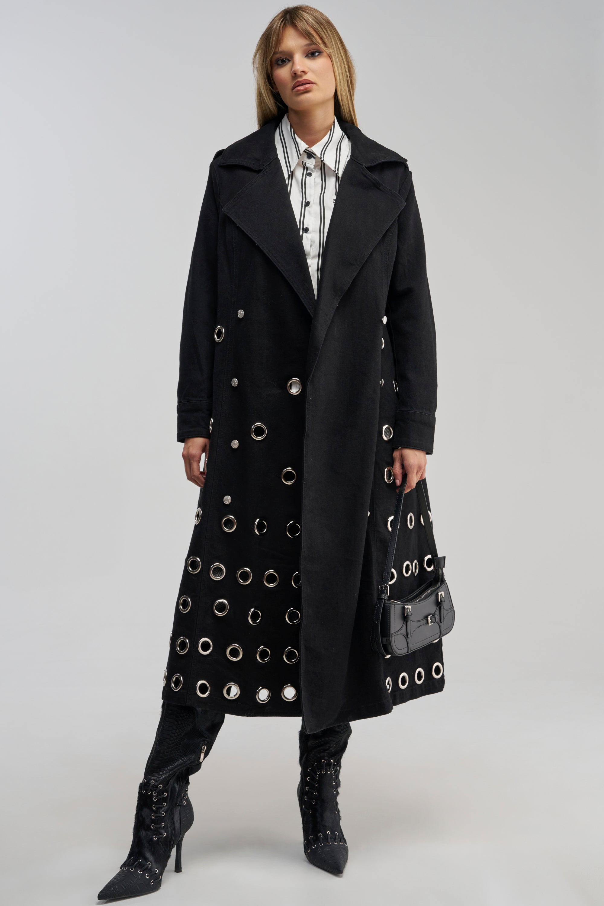 A woman wears the PHOENIX DENIM TRENCH—a long black denim coat with silver grommet details—paired with black lace-up boots and a white button-up shirt, carrying a small black handbag. She embodies modern streetwear style against a light gray background.