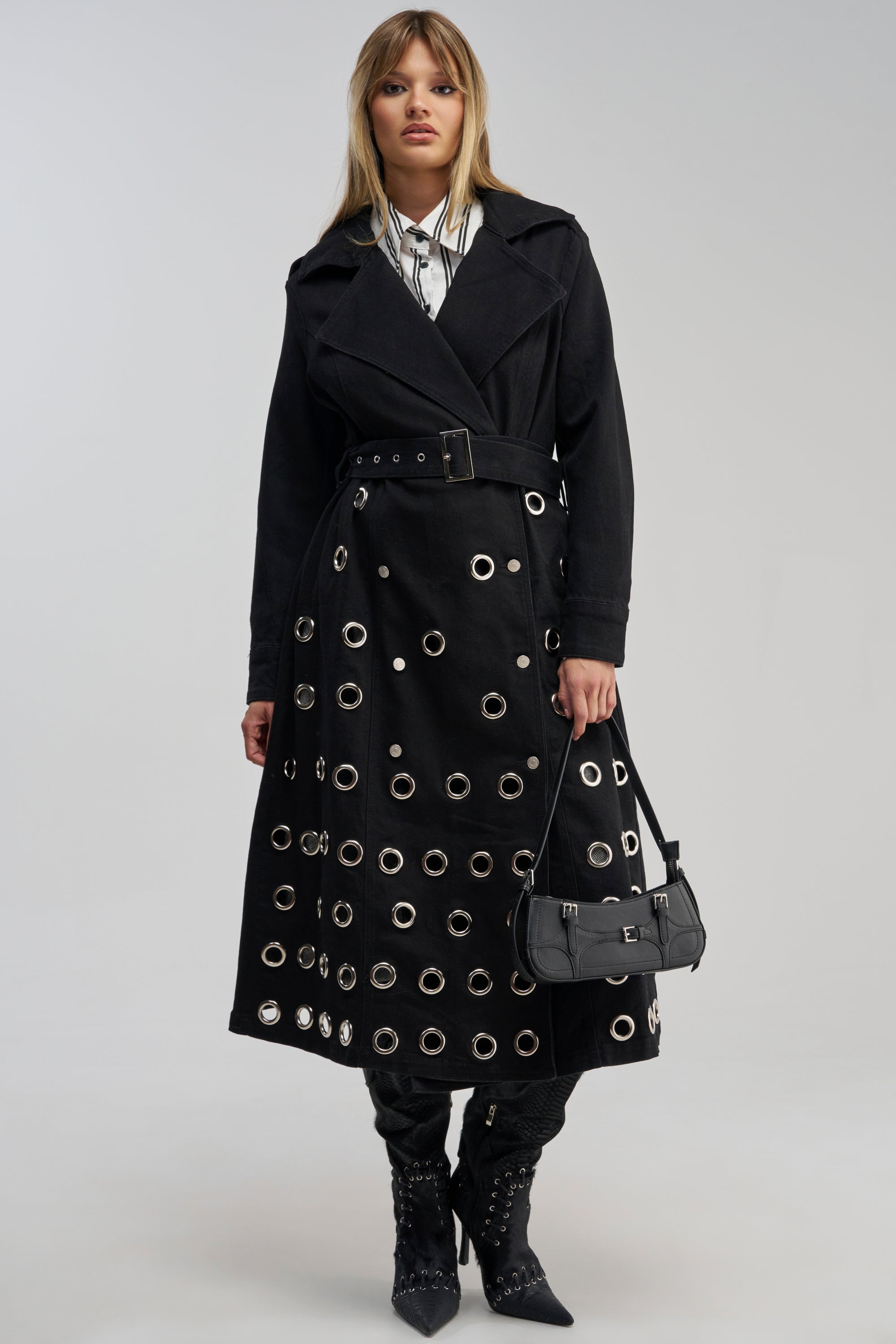 A woman faces forward against a plain background, embodying effortless streetwear style in the PHOENIX DENIM TRENCH—a black denim trench coat with silver grommets—layered over a collared shirt, paired with black boots and a black handbag.