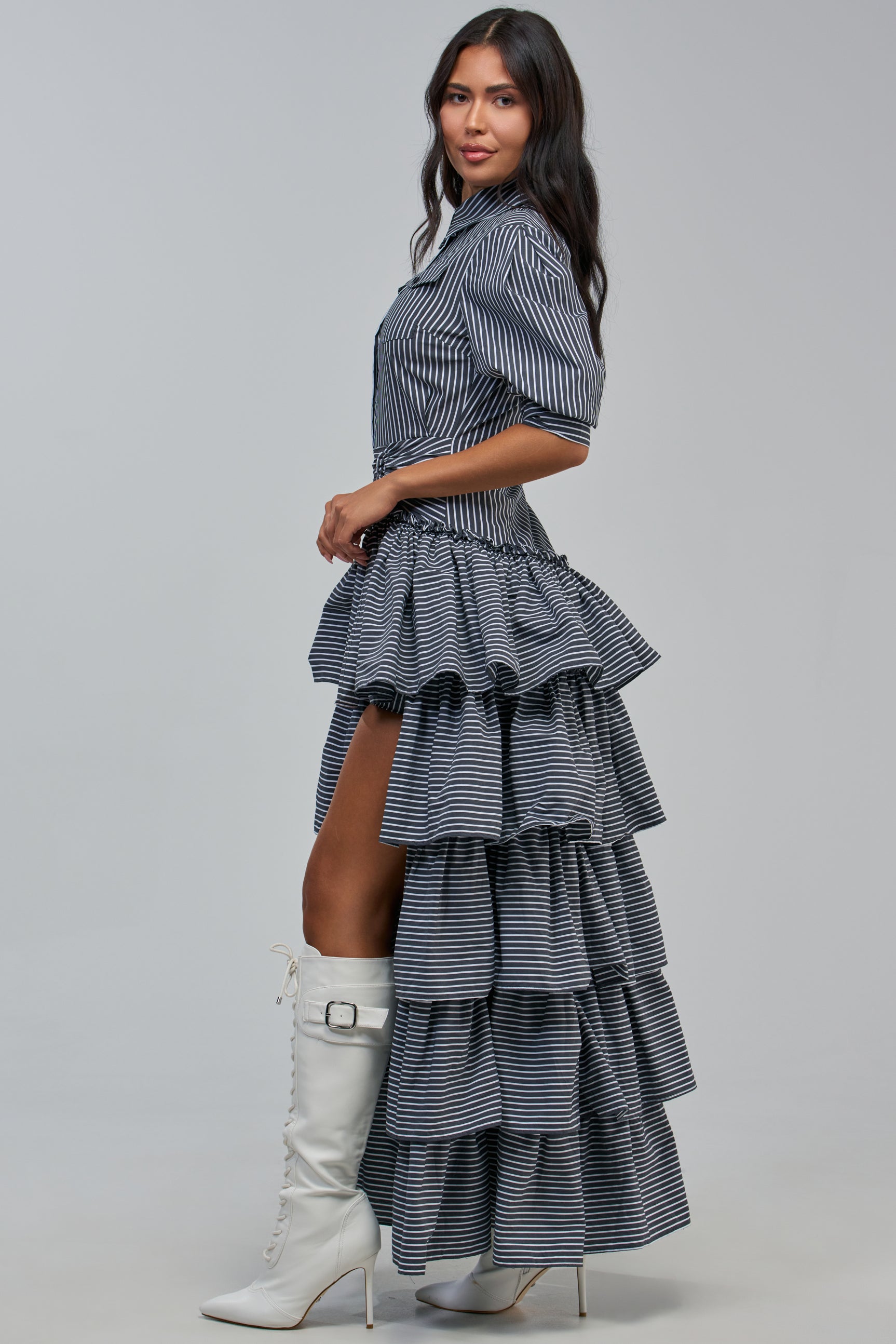 A woman wearing the IT’S A DATE POPLIN MAXI DRESS IN GREY PINSTRIPE—featuring blue and white stripes, ruffled high-low hem, and a silhouette reminiscent of a maxi dress—stands sideways against a plain light background, paired with white heeled knee-high boots.