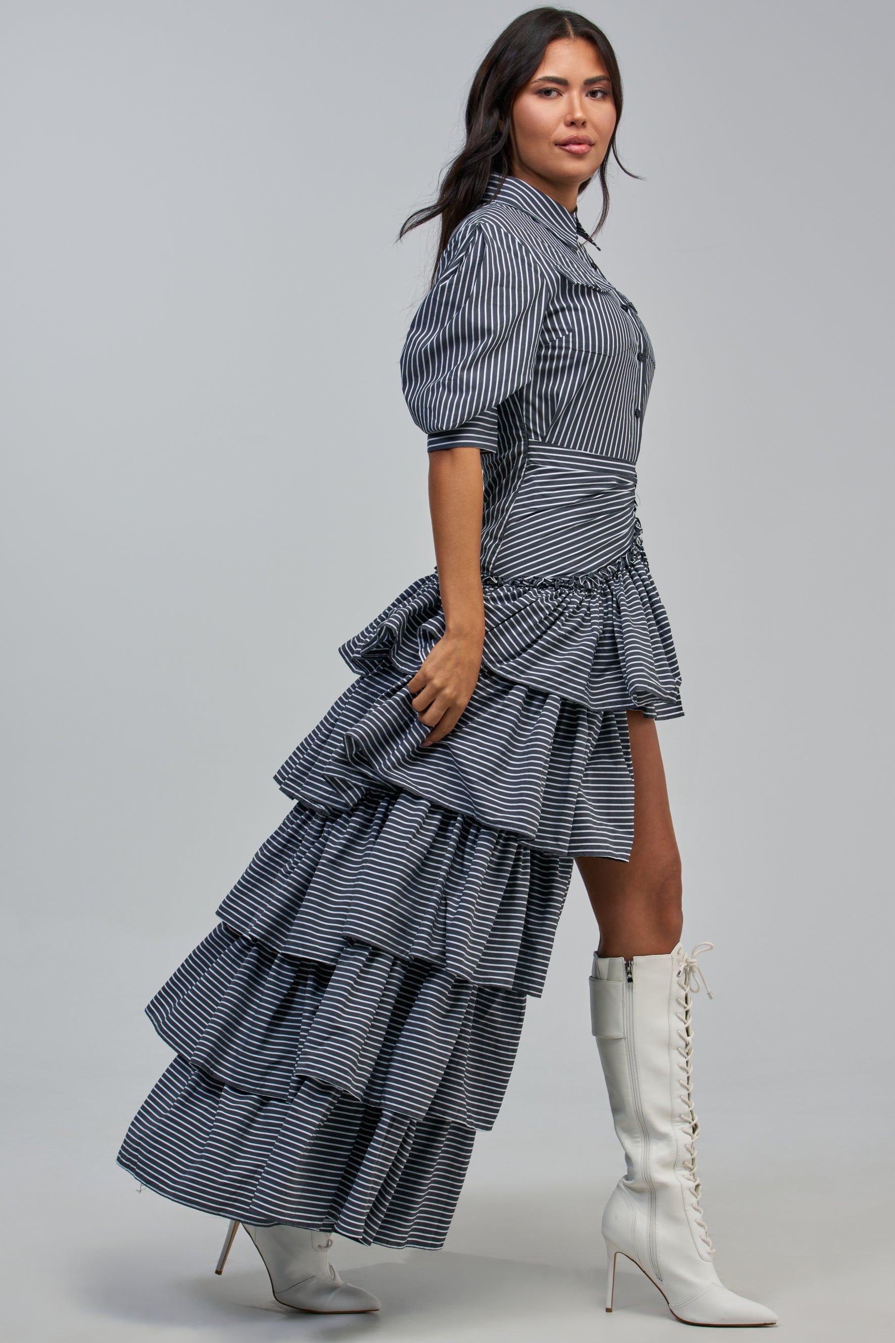 A woman wearing the IT’S A DATE POPLIN MAXI DRESS IN GREY PINSTRIPE, featuring black and white stripes with layered ruffles that are high in the front and long in the back, stands sideways. She pairs the maxi dress with knee-high white lace-up boots and has long dark hair styled in loose waves.