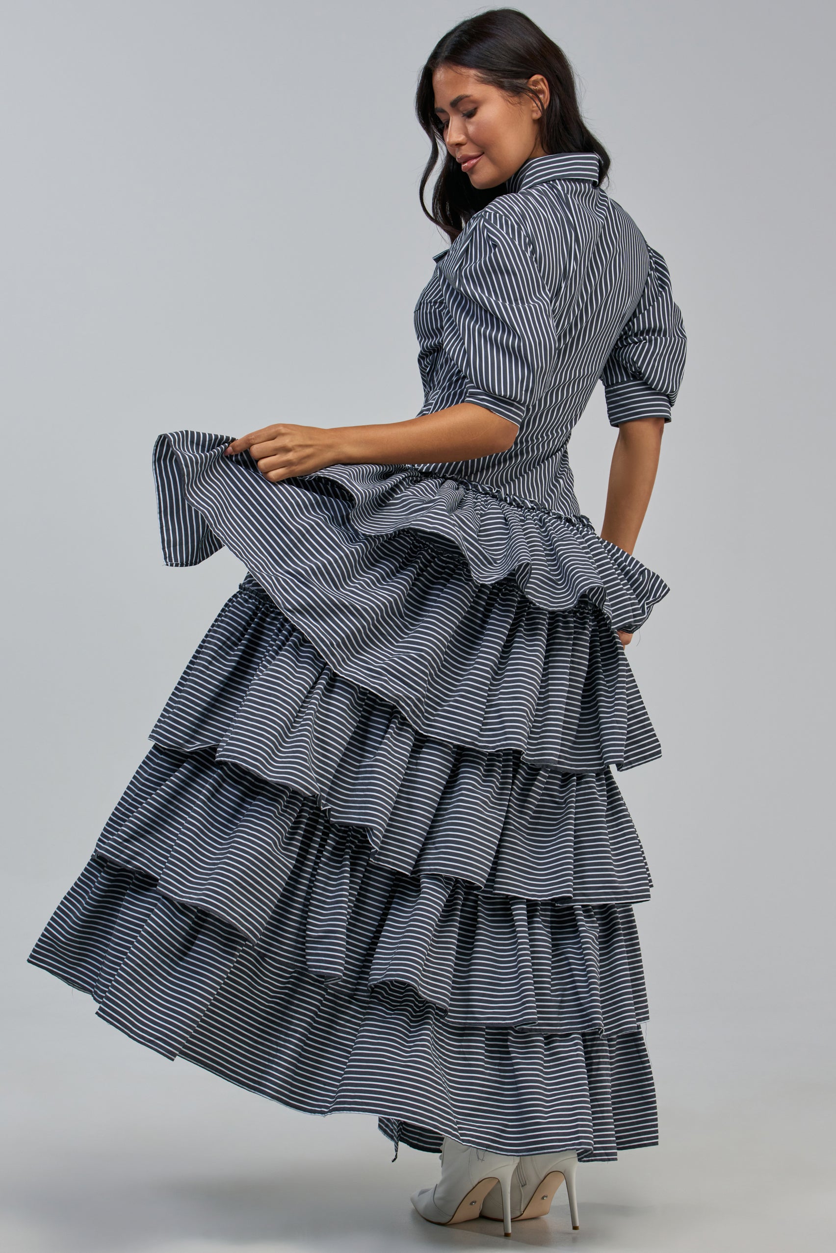 A woman poses sideways, lifting a layer of her skirt while wearing the IT’S A DATE POPLIN MAXI DRESS IN GREY PINSTRIPE. She has long, dark hair and pairs the maxi dress with high-heeled shoes.