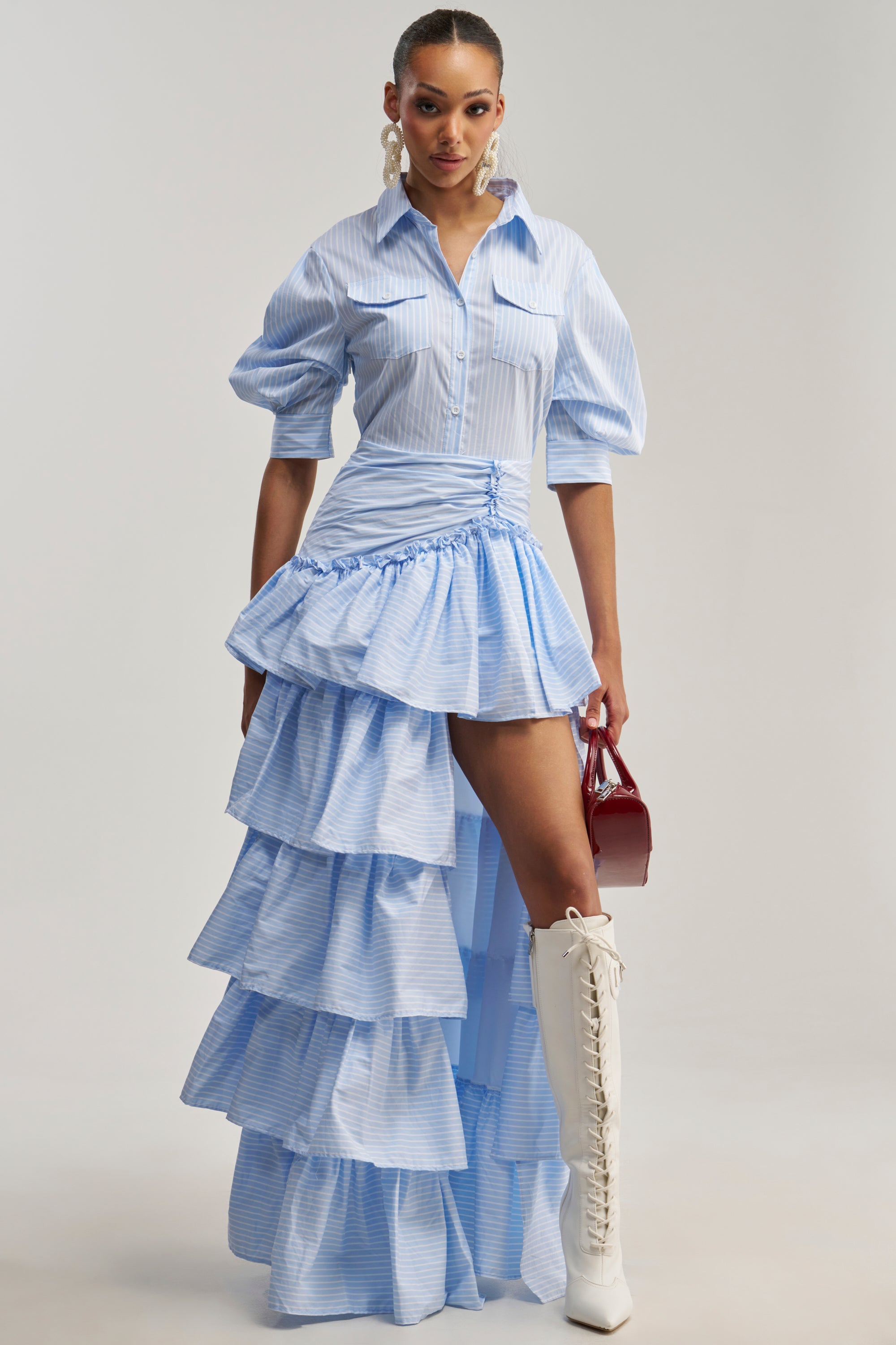 Wearing the IT’S A DATE POPLIN MAXI DRESS IN BLUE, featuring ruffled details and a high-low hem, paired with white lace-up knee boots, statement earrings, and a small red handbag, she stands against a plain light background—truly the perfect statement brunch look.